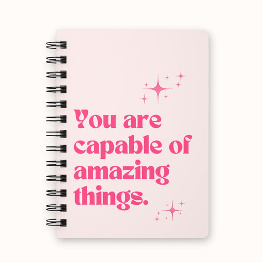 Light pink spiral notebook with 'You are capable of amazing things' text and sparkle details on front cover