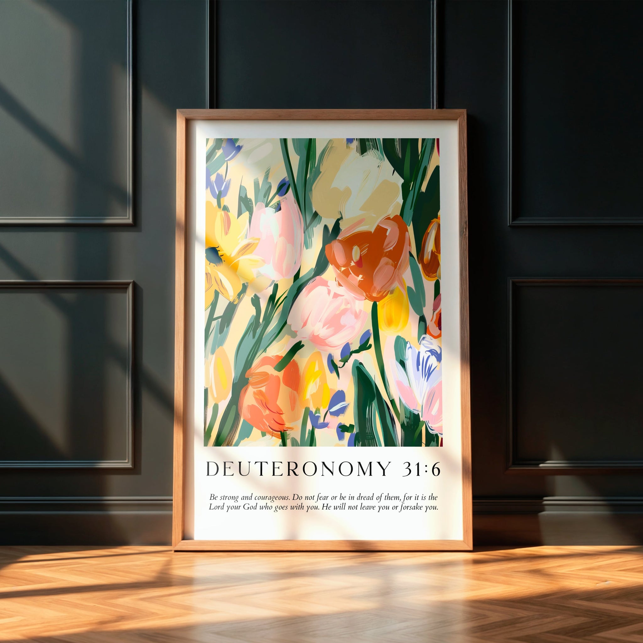 Modern Christian typography print with Deuteronomy 31:6 and soft florals. In a wooden frame leaning on a black panelled wall.