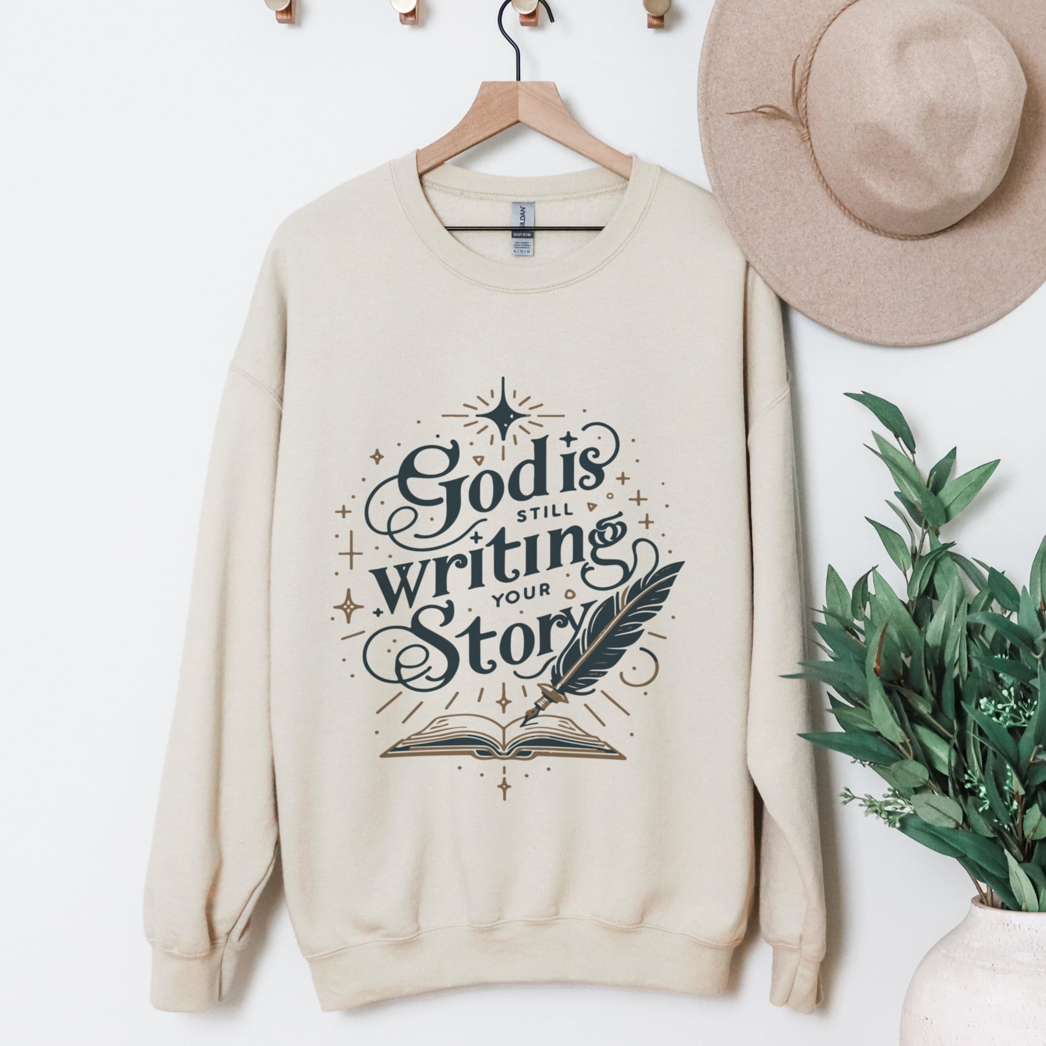 Beige God Is Still Writing Your Story sweatshirt hanging display - beautiful book design Christian crewneck clothing