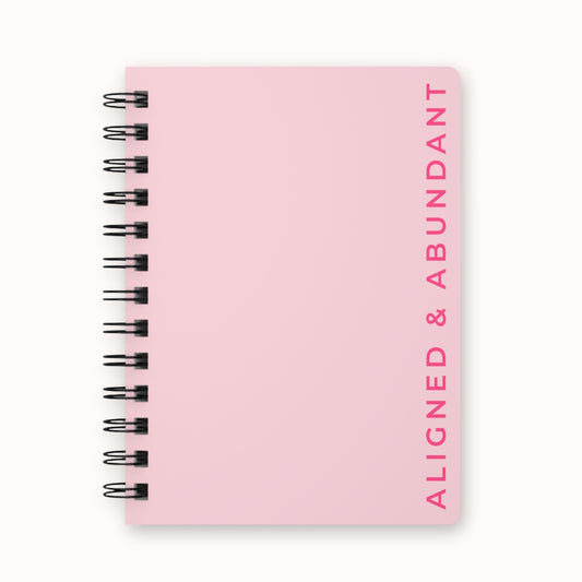 Light pink spiral notebook with 'Aligned & Abundant' text running vertically down the front cover
