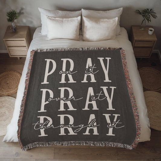 A soft, dark gray woven blanket with the words "Pray on it. Pray over it. Pray through it" stitched in elegant lettering, framed with a colorful fringe.