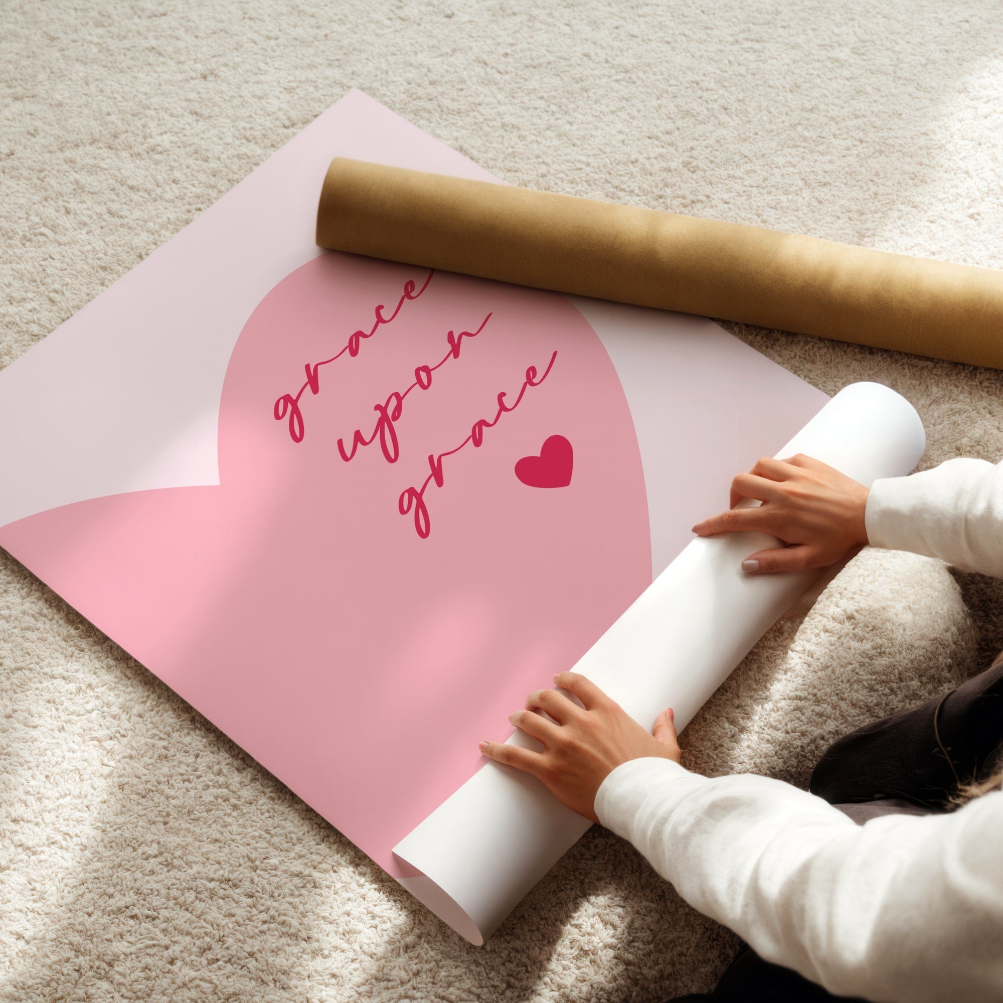 Person unrolling pink poster with 'grace upon grace' text and a heart on a light-colored floor.