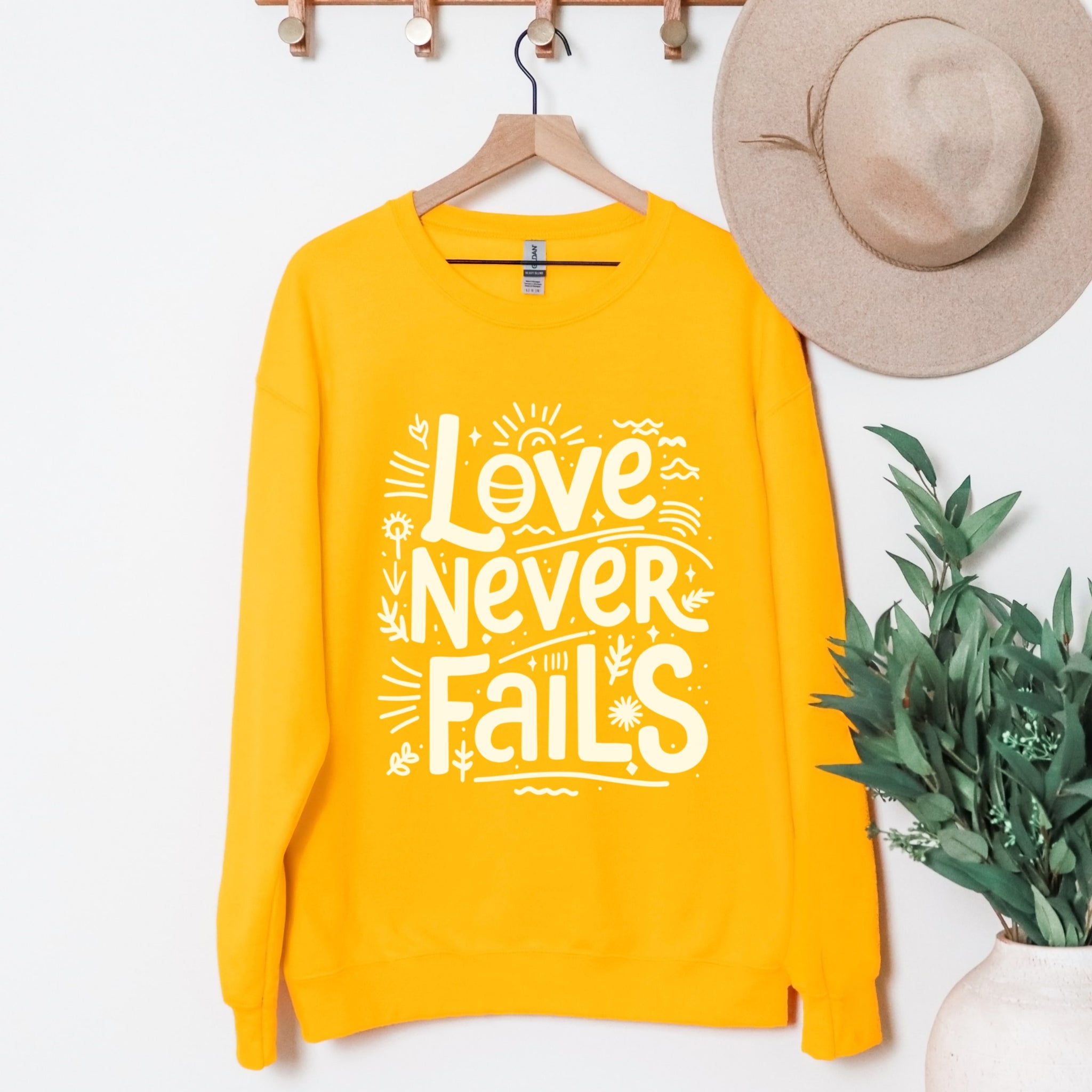 Love Never Fails yellow sweatshirt hanging display with playful lettering - positive Christian faith crewneck for women