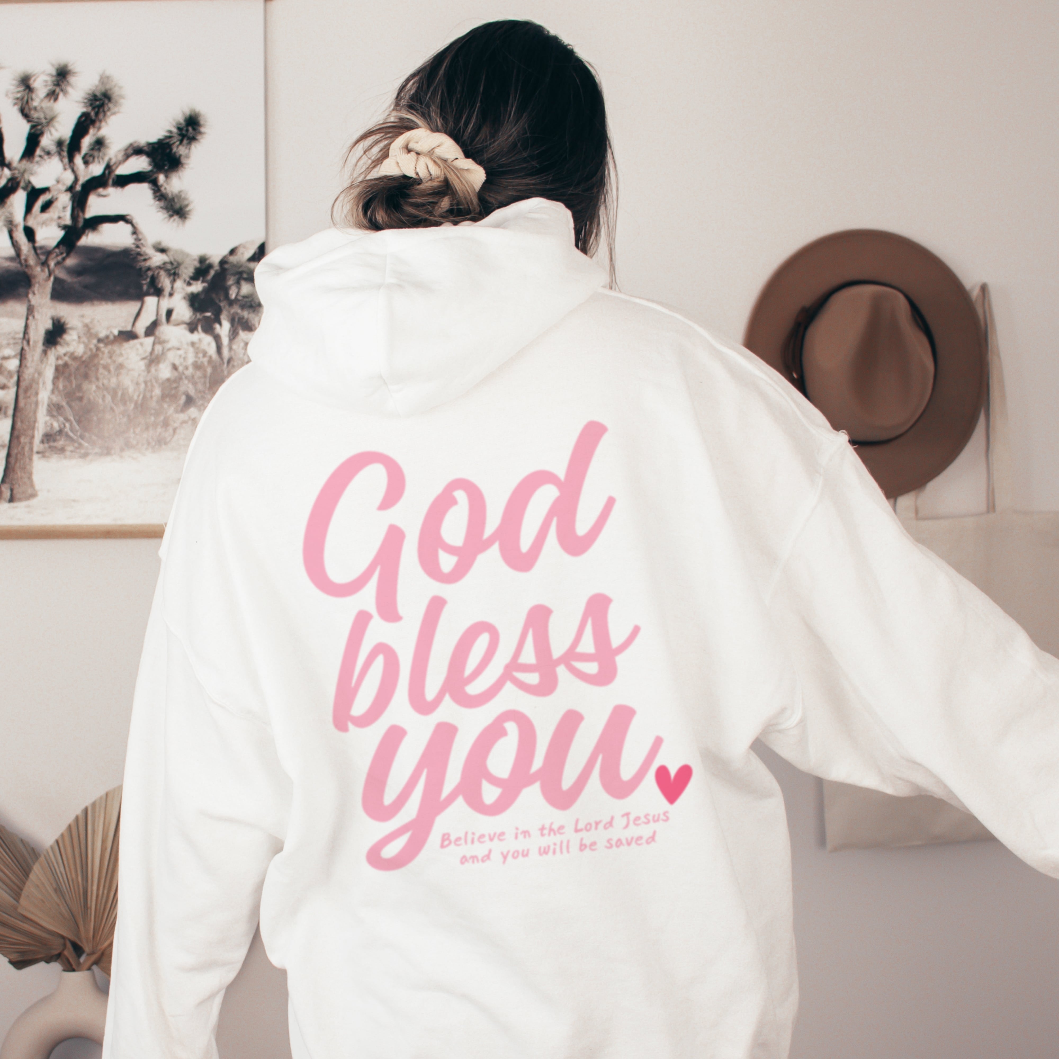 Back of white Christian hoodie with pink God Bless You lettering and scripture verse believe in the Lord Jesus, faith-based clothing for women