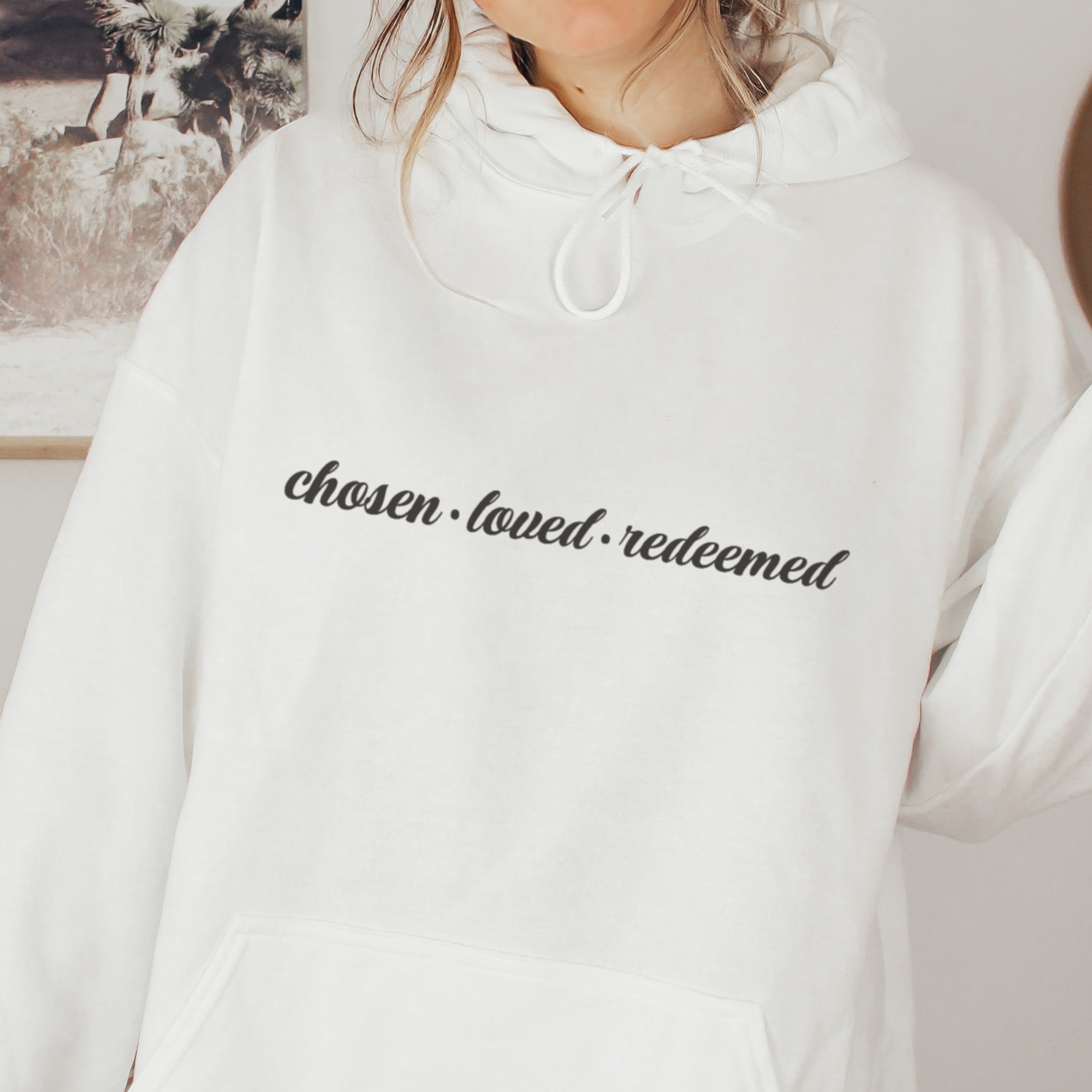 White hoodie with 'chosen loved redeemed' text worn by a person indoors.