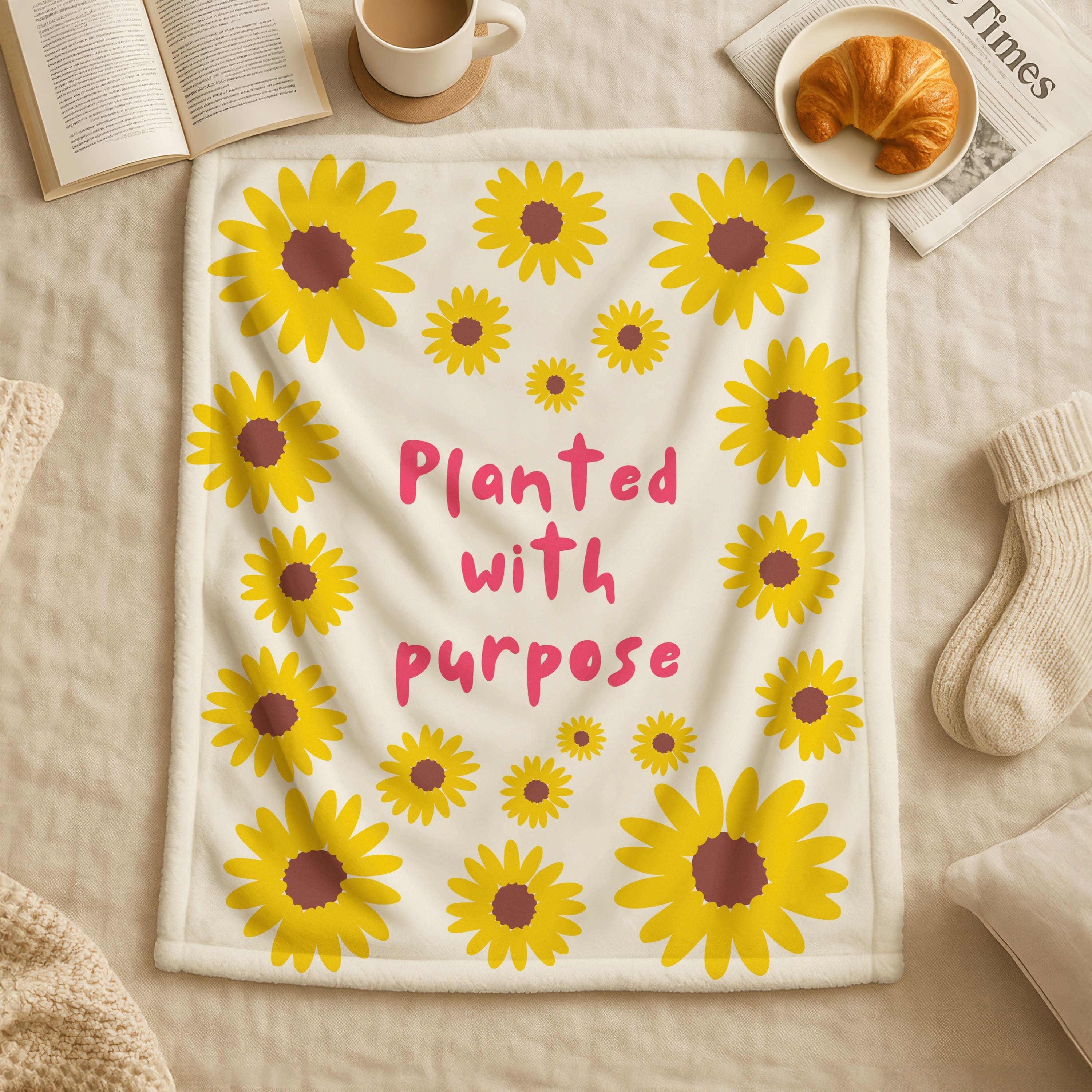 Blanket with sunflower pattern and 'Planted with purpose' text on a cozy setting with coffee, croissant, and book.