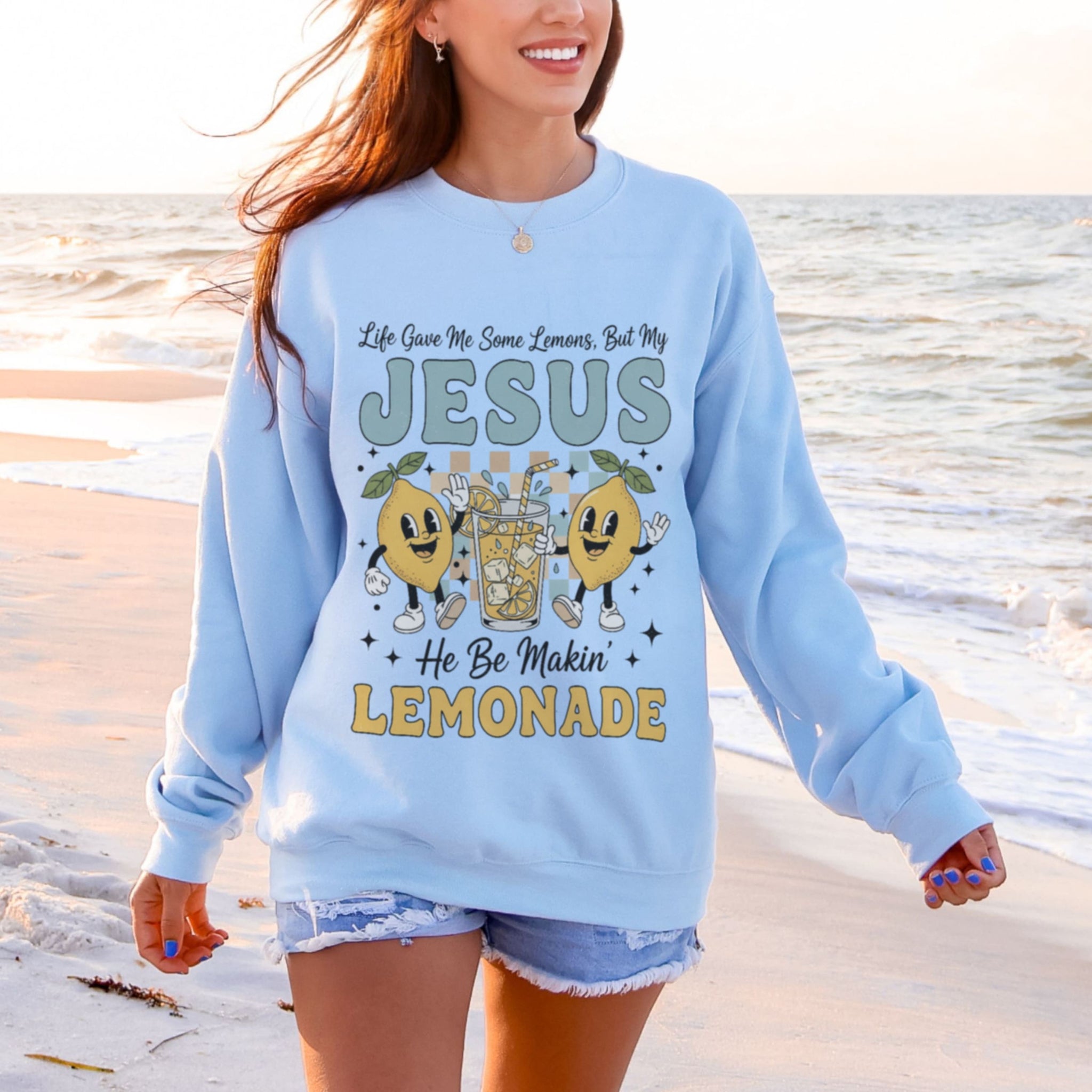Woman wearing light blue Jesus Making Lemonade sweatshirt at beach - fun Christian crewneck with lemon design for women