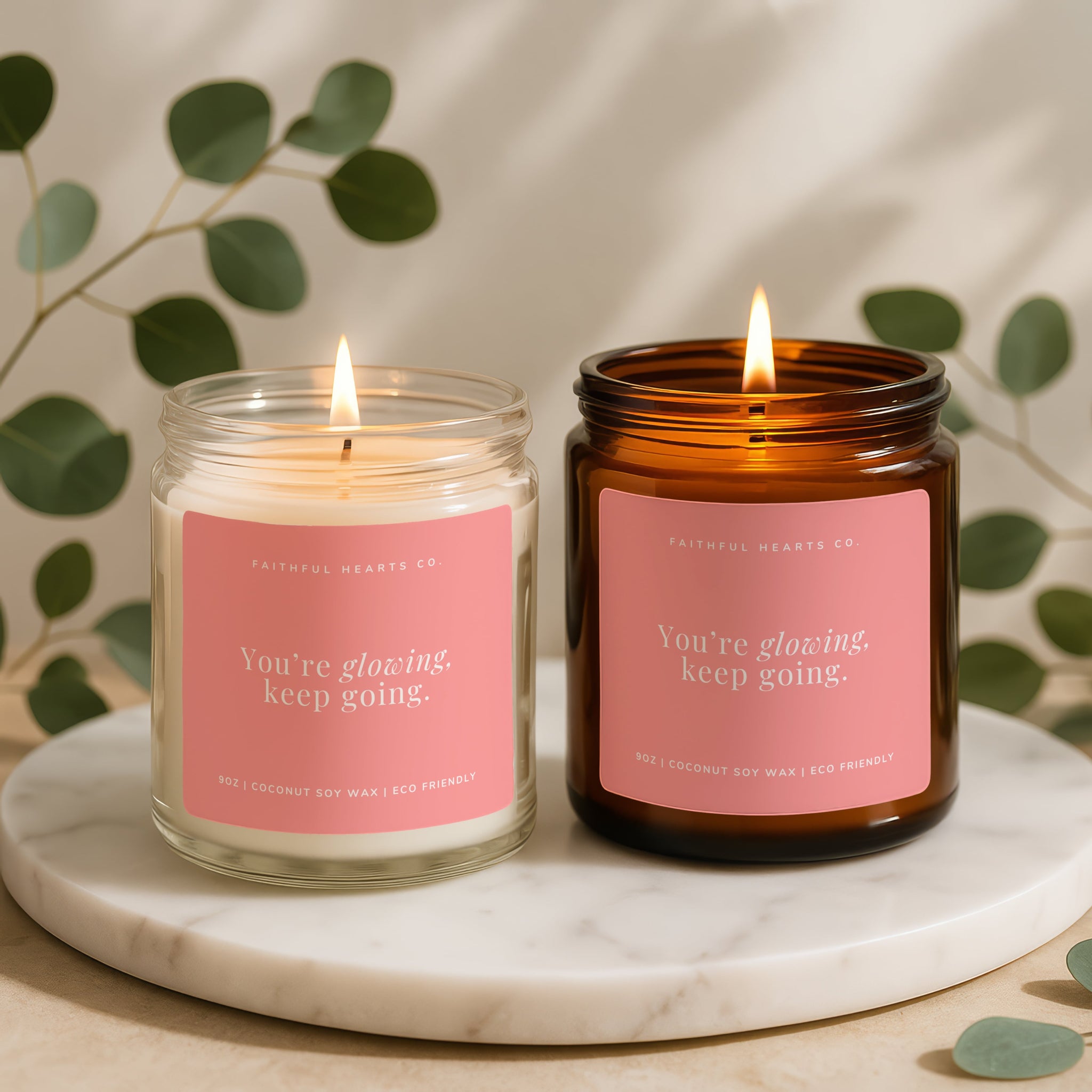 Two candles with pink labels on a marble surface with green leaves in the background