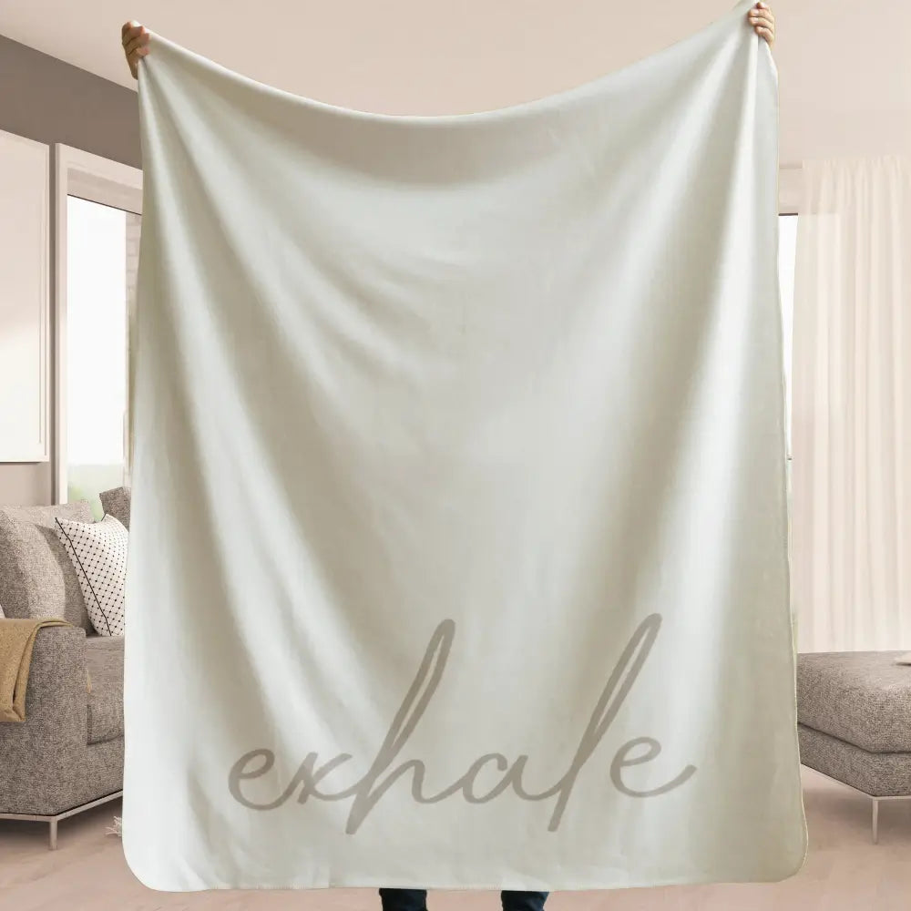 Neutral colored sherpa fleece blanket with exhale written on the front being held up to show how large it is