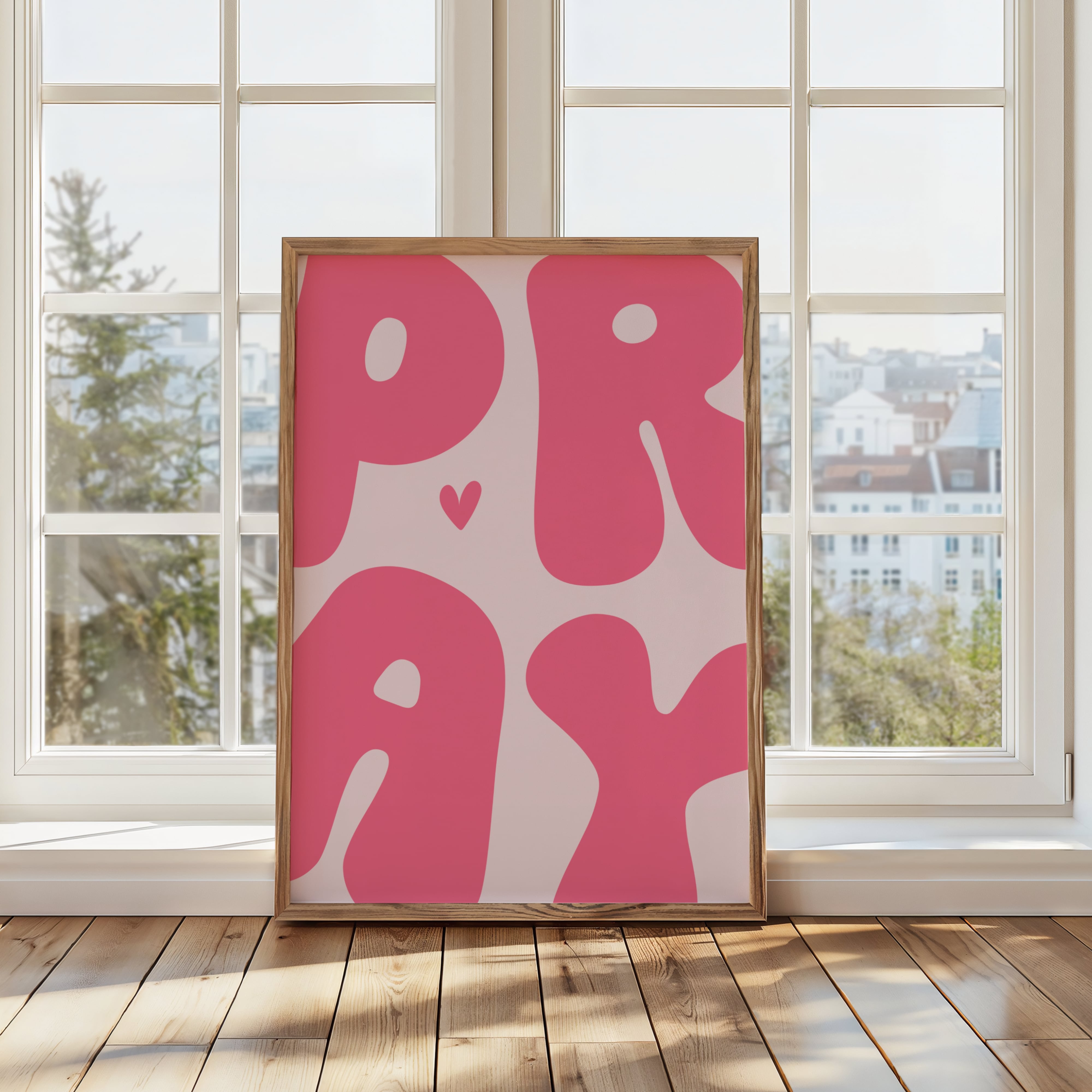 Framed artwork with 'PRAY' in pink bubble letters on a wall in a room with large windows.