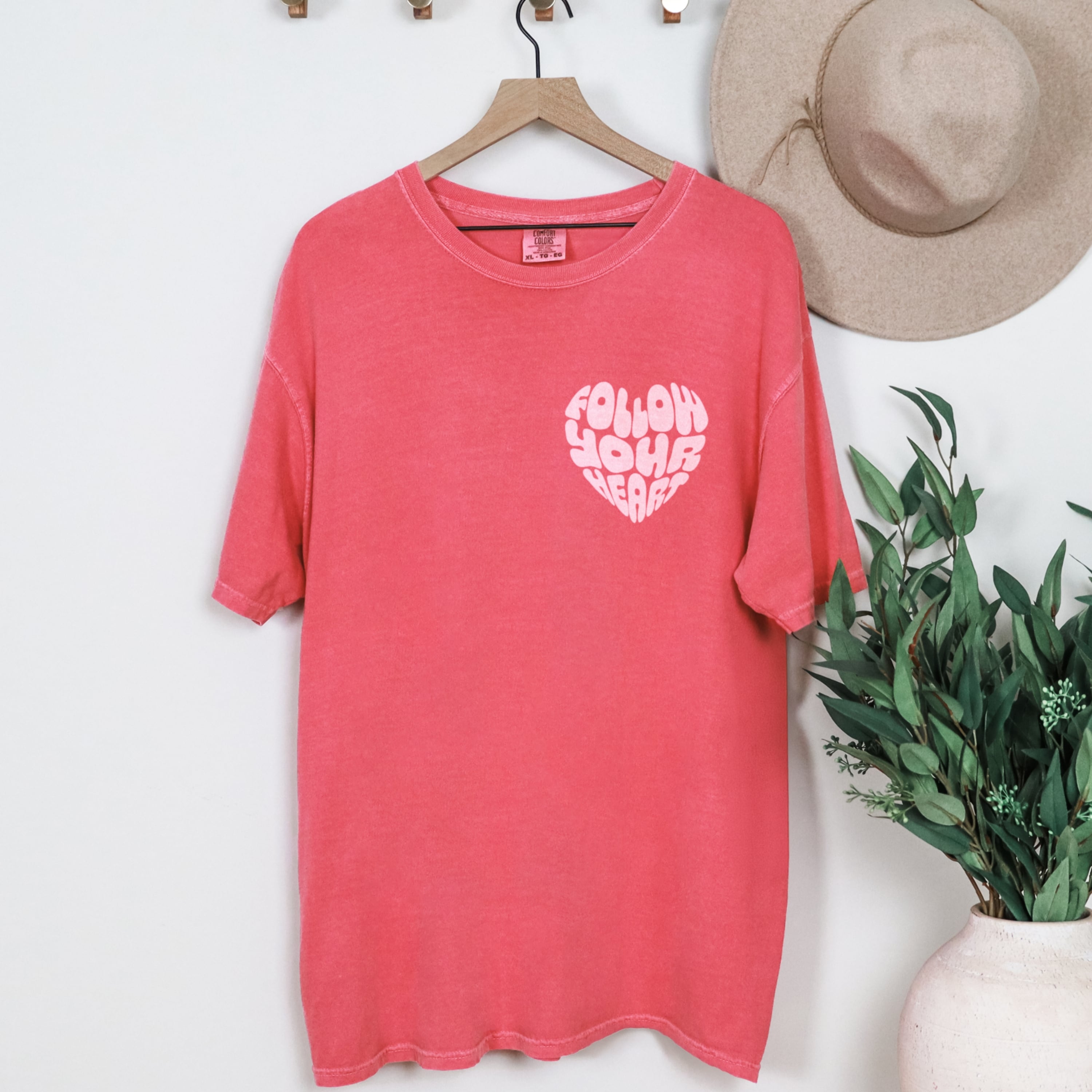Coral pink t-shirt with 'Follow Your Heart' design on a hanger with a hat and plant in the background.