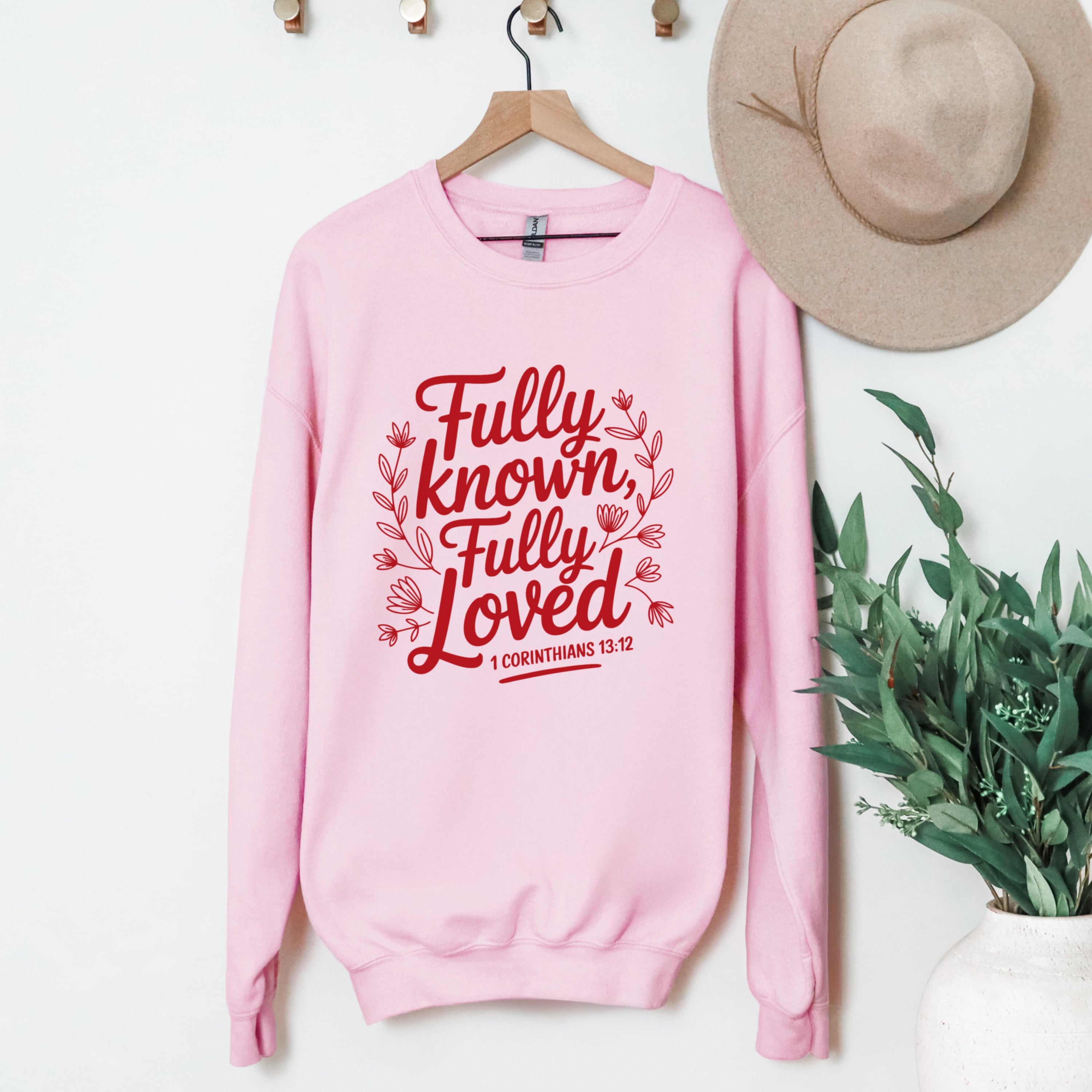 Pink sweatshirt with red text 'Fully known, Fully Loved' on a hanger with a hat and plant in the background.
