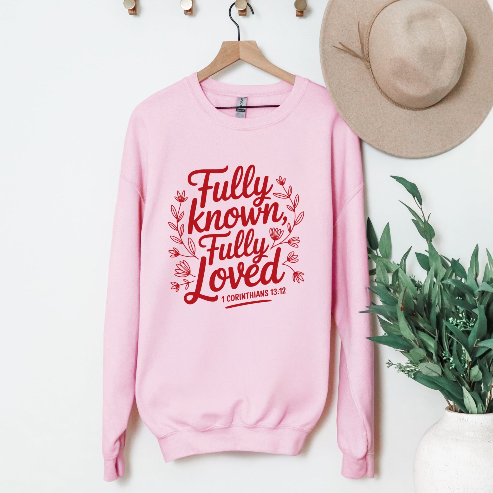 Pink sweatshirt with red text 'Fully known, Fully Loved' on a hanger with a hat and plant in the background.