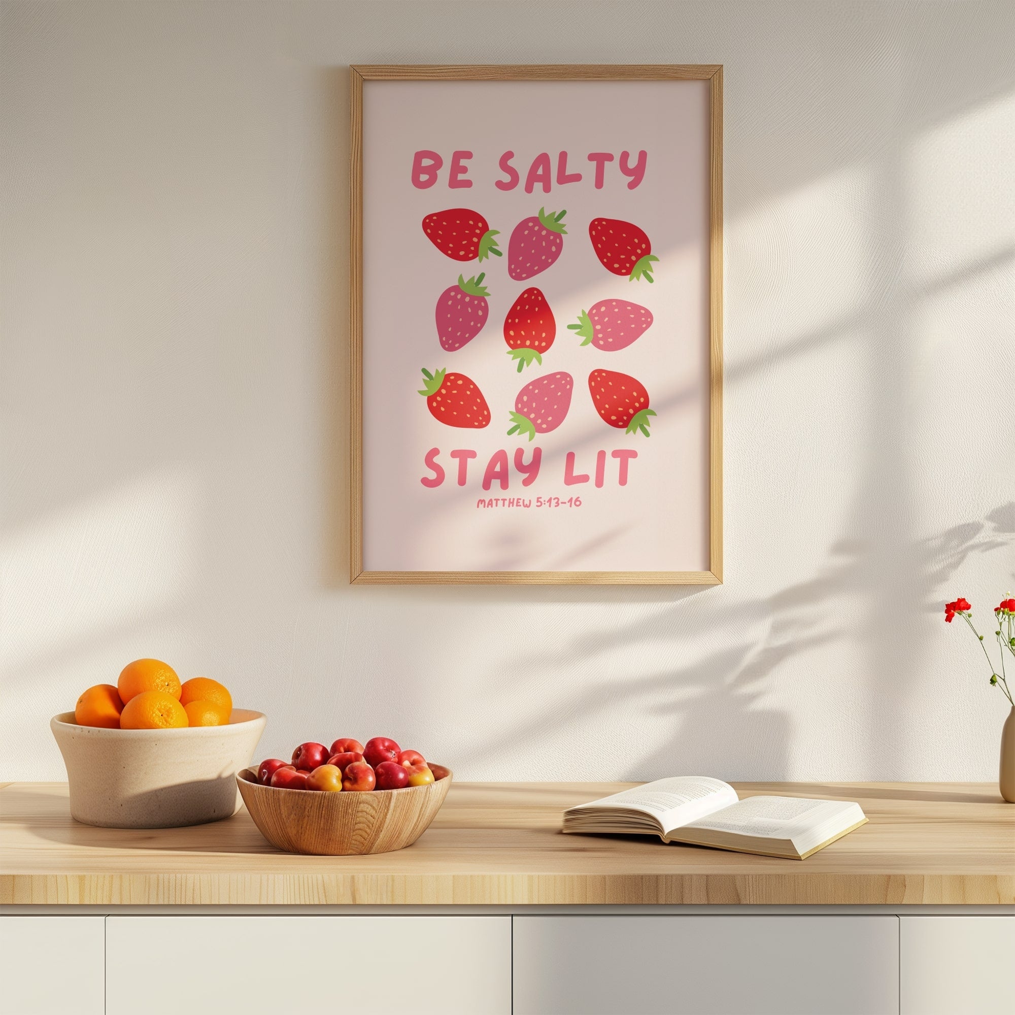Framed artwork with strawberry design and scripture on a wall above a wooden table with fruit and a book.