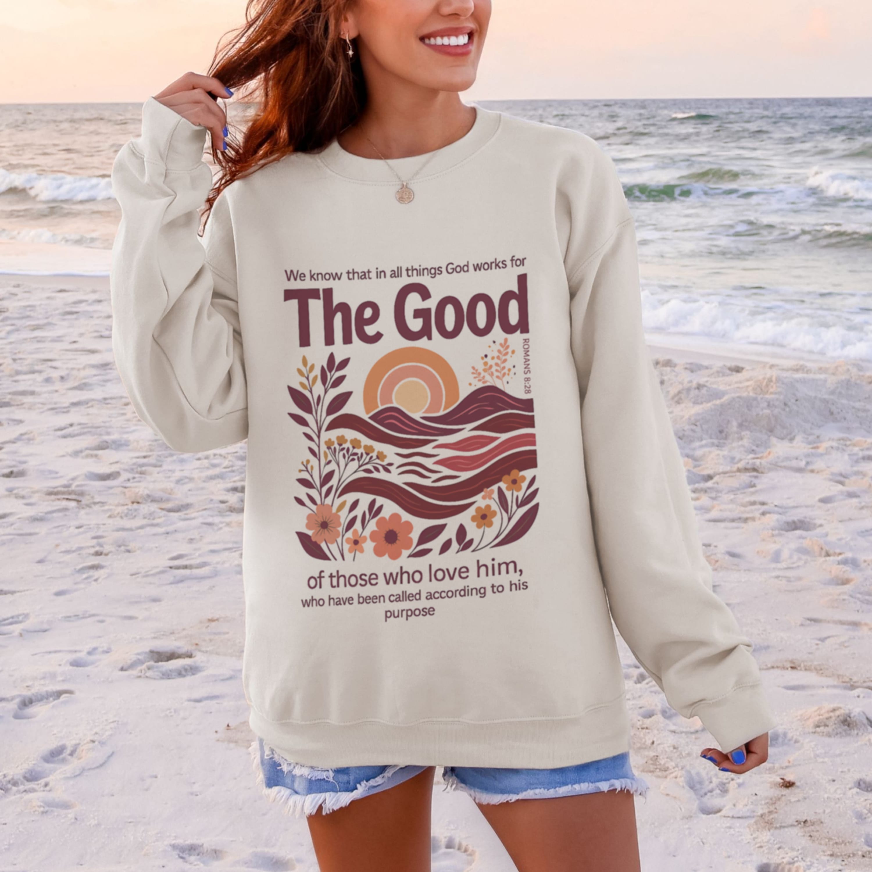 Woman wearing beige God Works For The Good Romans 8:28 sweatshirt at beach - boho Christian crewneck with sunset and wave design for women