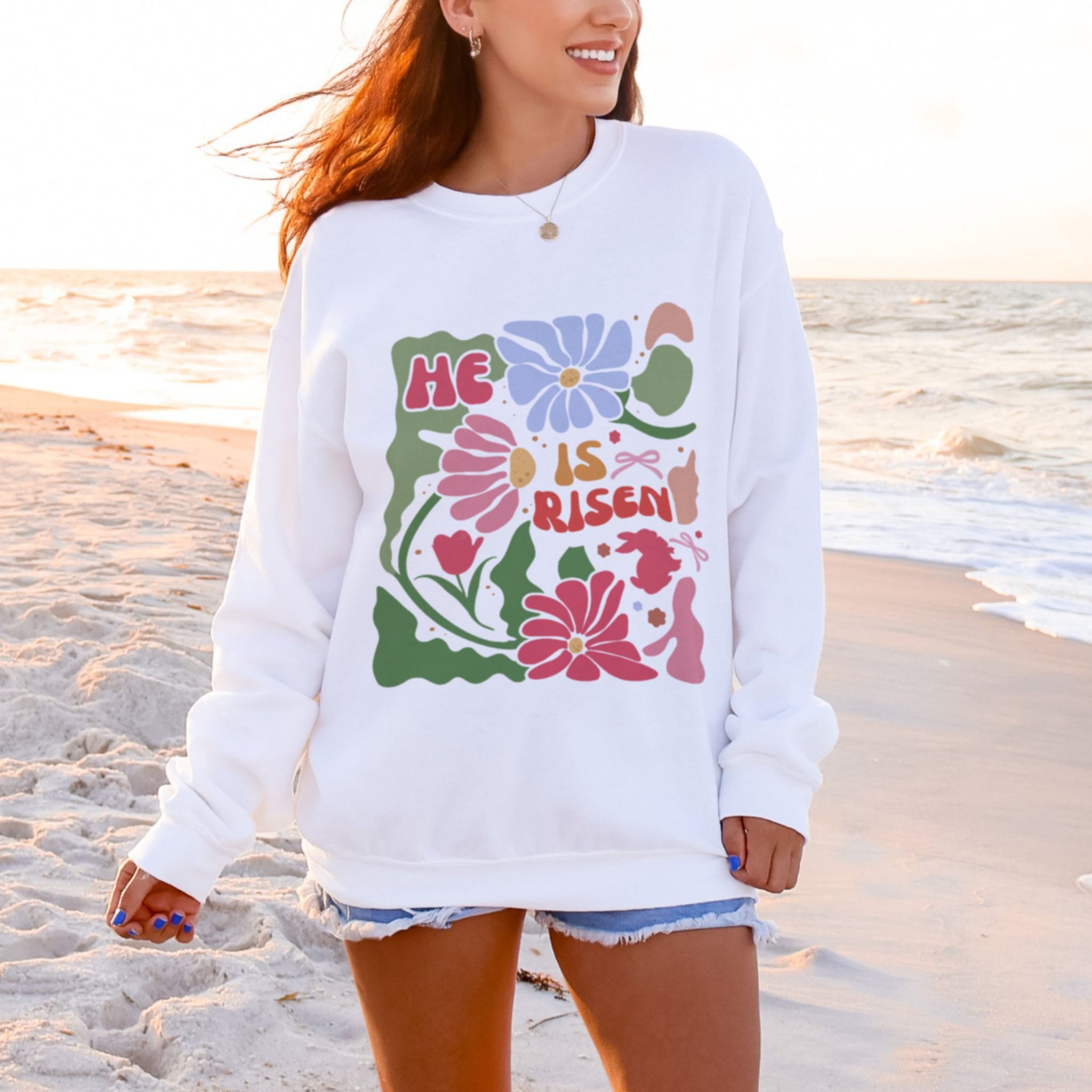 Woman wearing white He Is Risen sweatshirt with retro floral design at beach - Christian crewneck for women