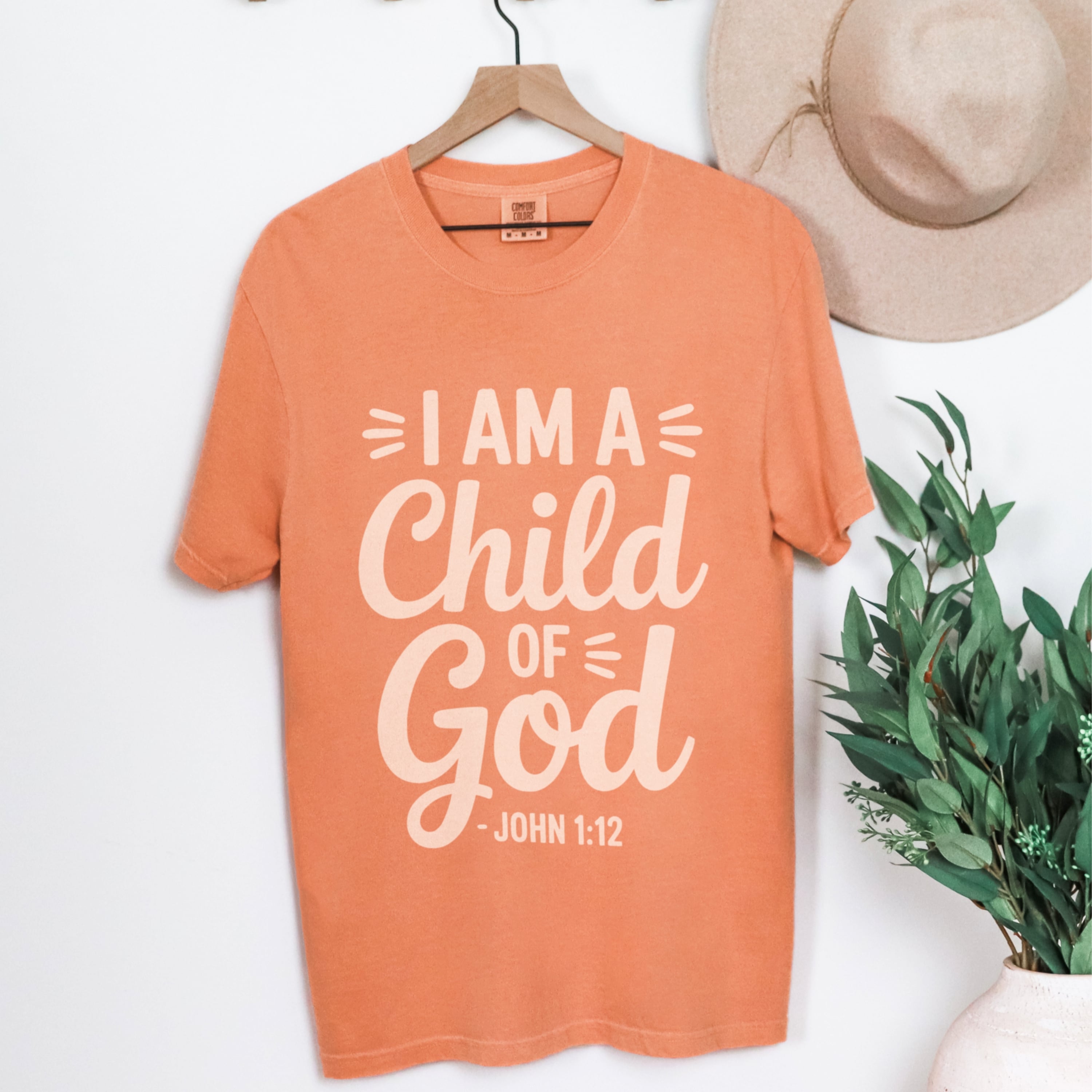 Close up of I Am A Child Of God design with John 1:12 scripture reference in cream on terracotta cotton tshirt