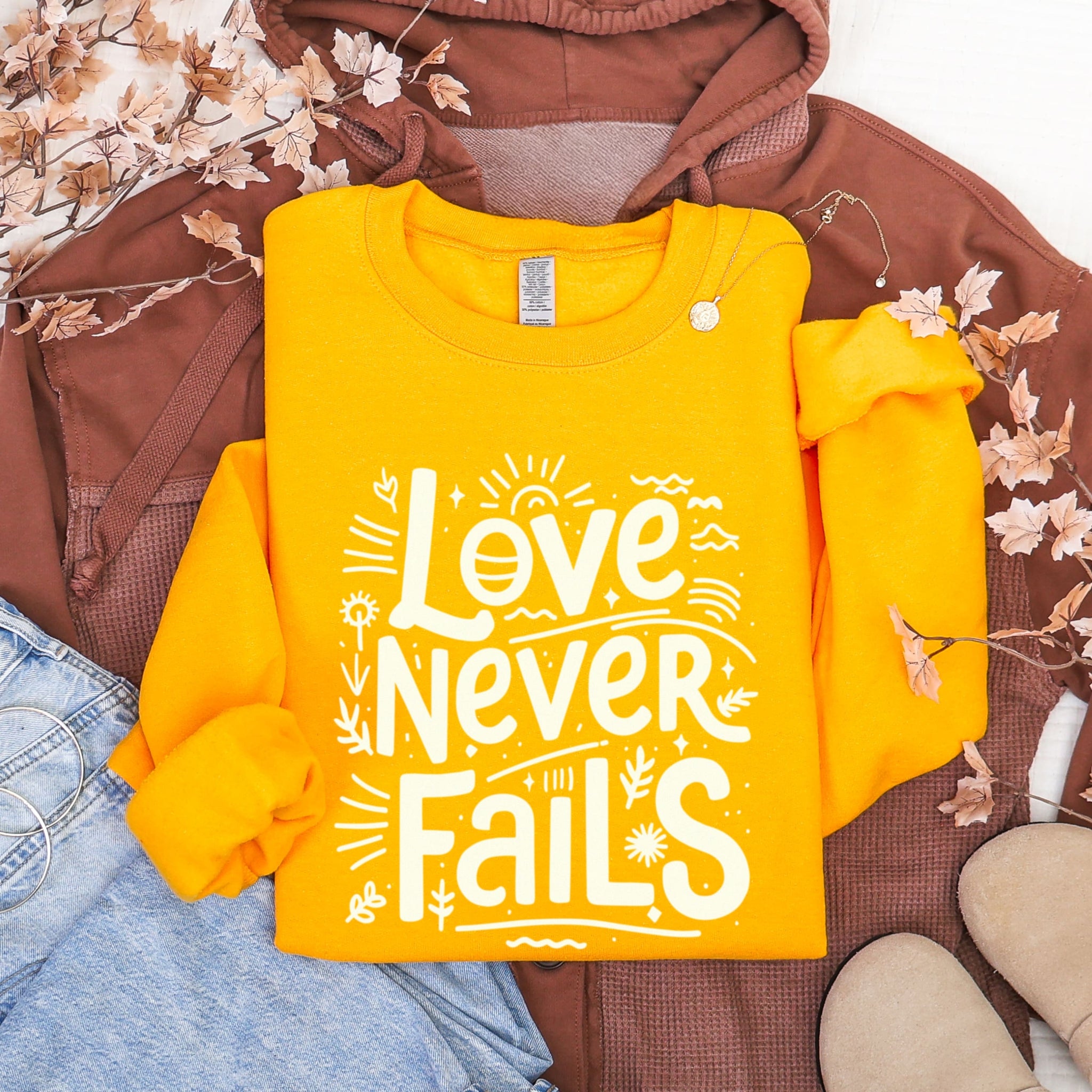 Bright yellow Love Never Fails sweatshirt flat lay showing detailed typography - 1 Corinthians 13:8 Christian apparel