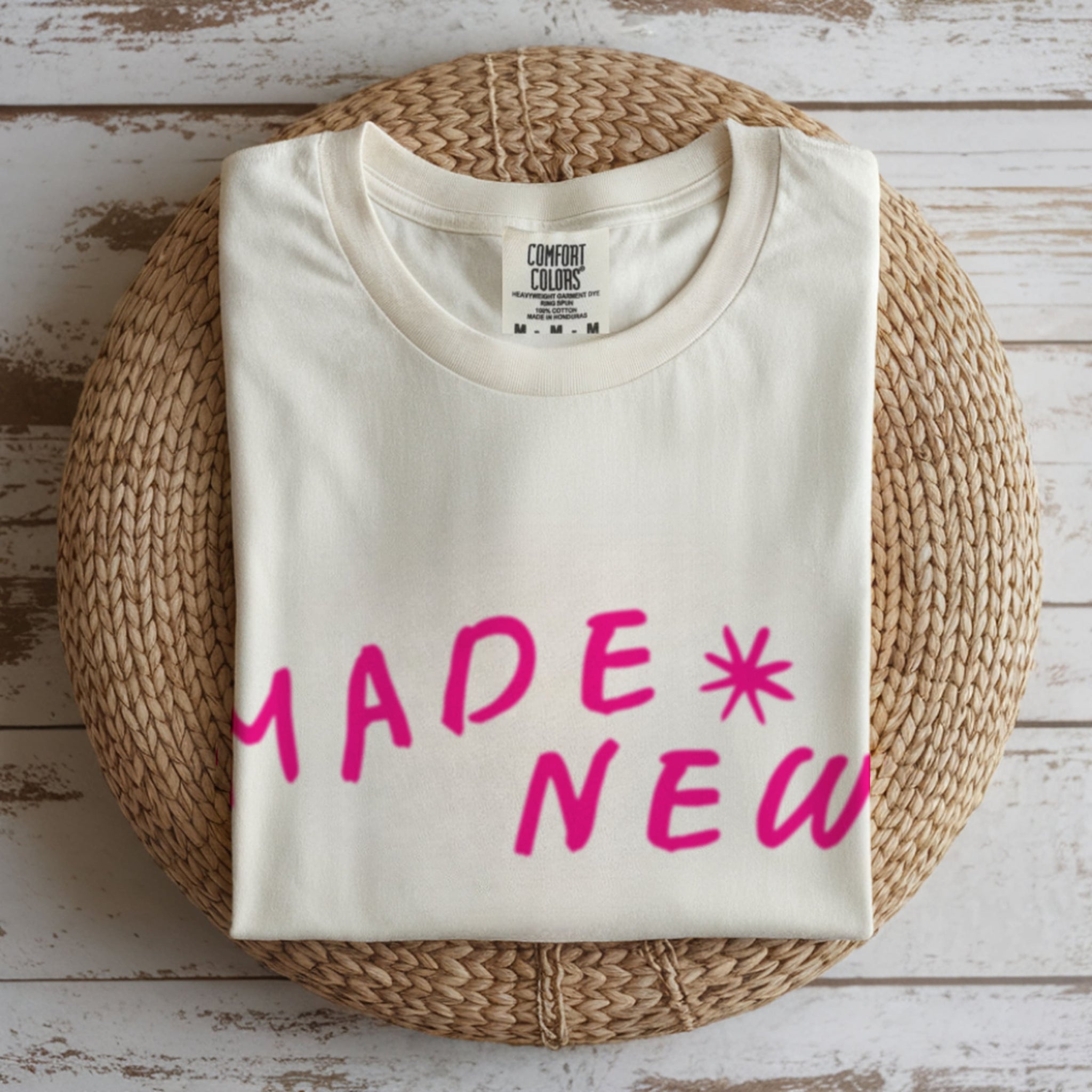 Folded cream t-shirt with pink text 'MADE NEW' on a woven mat.