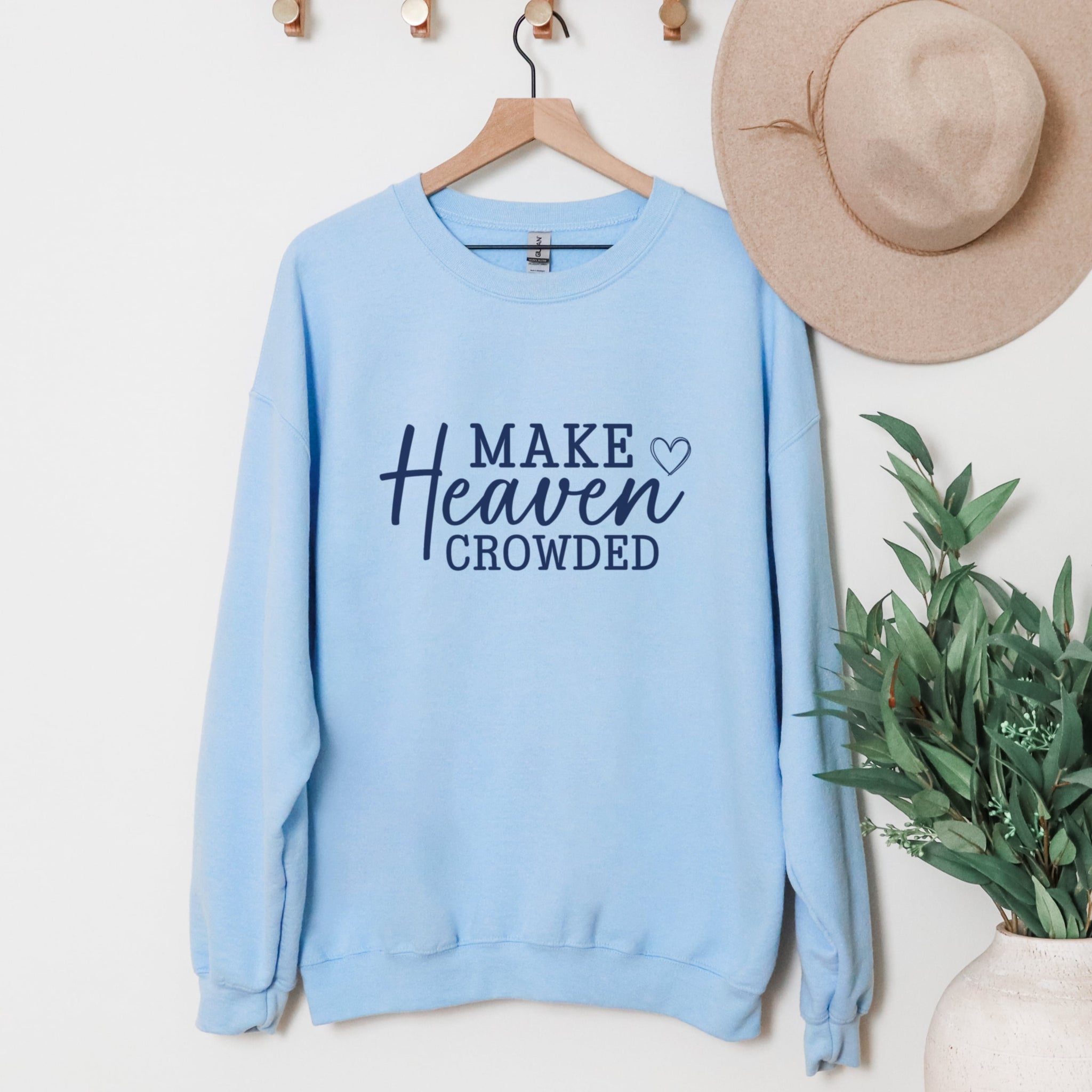 Light blue sweatshirt with 'MAKE HEAVEN CROWDED' text on a hanger with a hat and plant in the background.