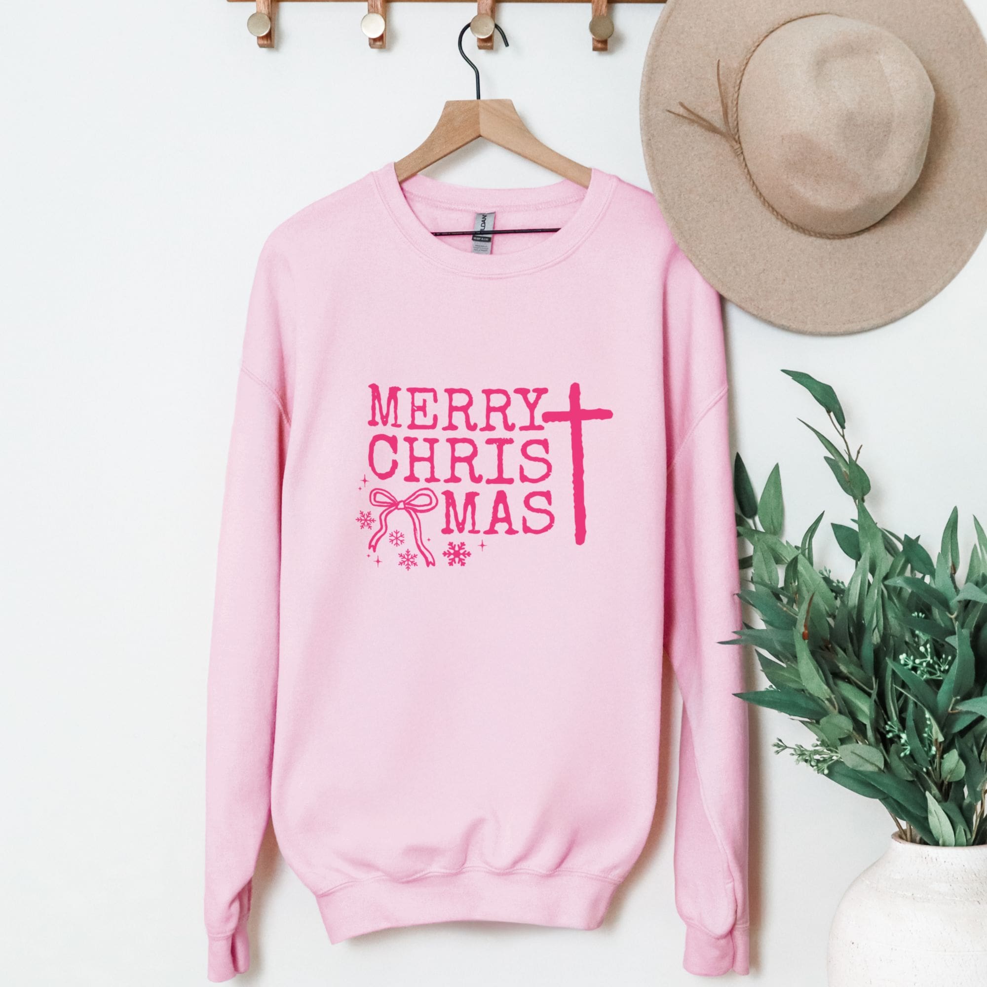 Pink sweatshirt with 'Merry Christmas' text on a hanger with a hat and plant in the background.