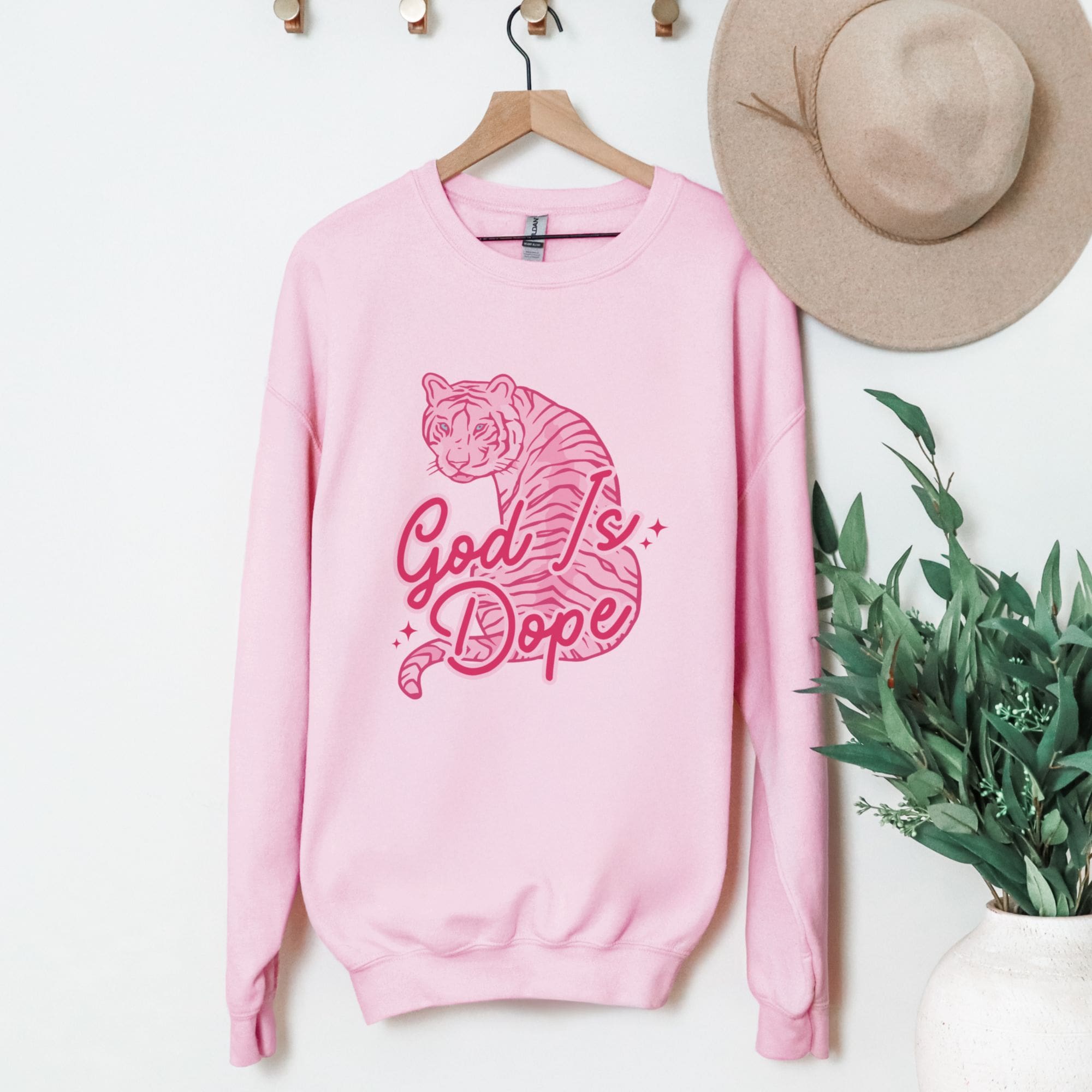 Pink sweatshirt with tiger graphic and 'God is Dope' text on a hanger with a hat and plant in the background.