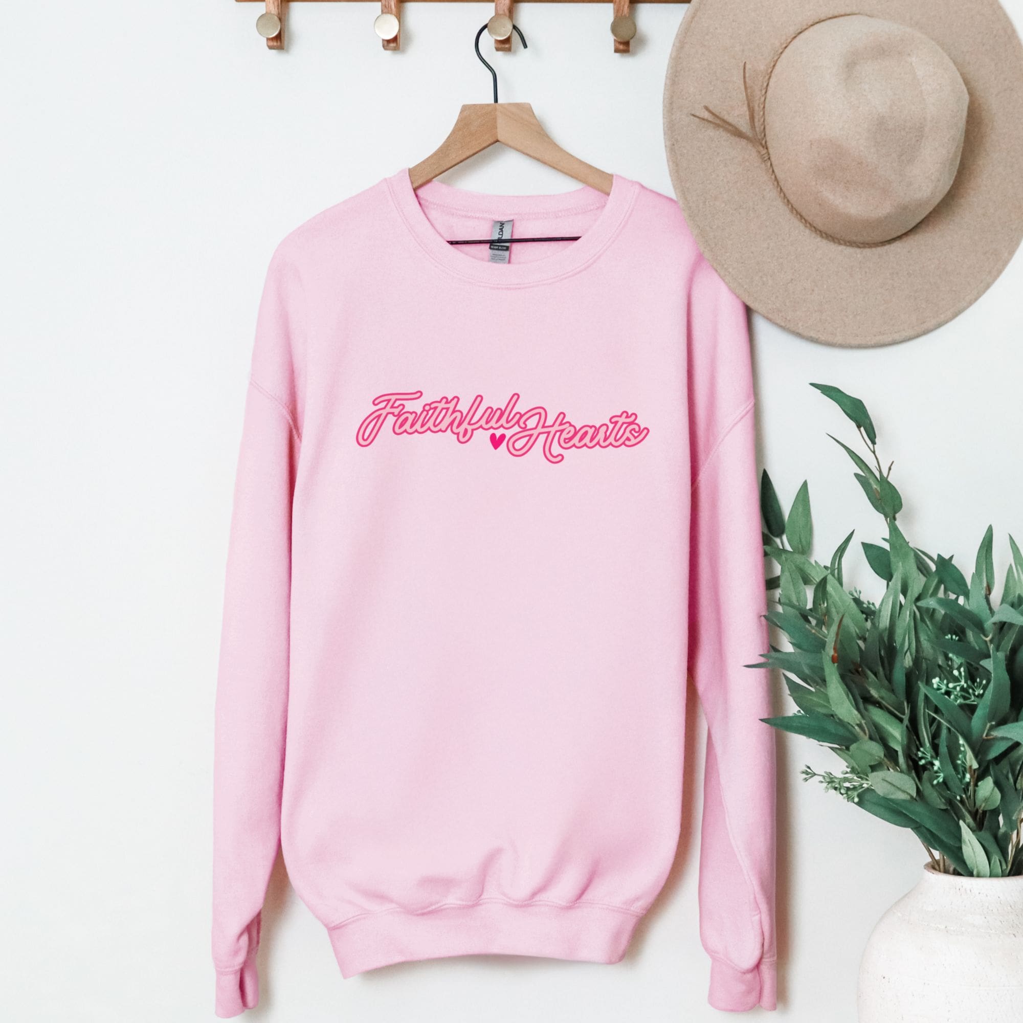 Pink Faithful Hearts crewneck sweatshirt hanging on wooden hanger with beige hat - cute Christian faith-based apparel for women