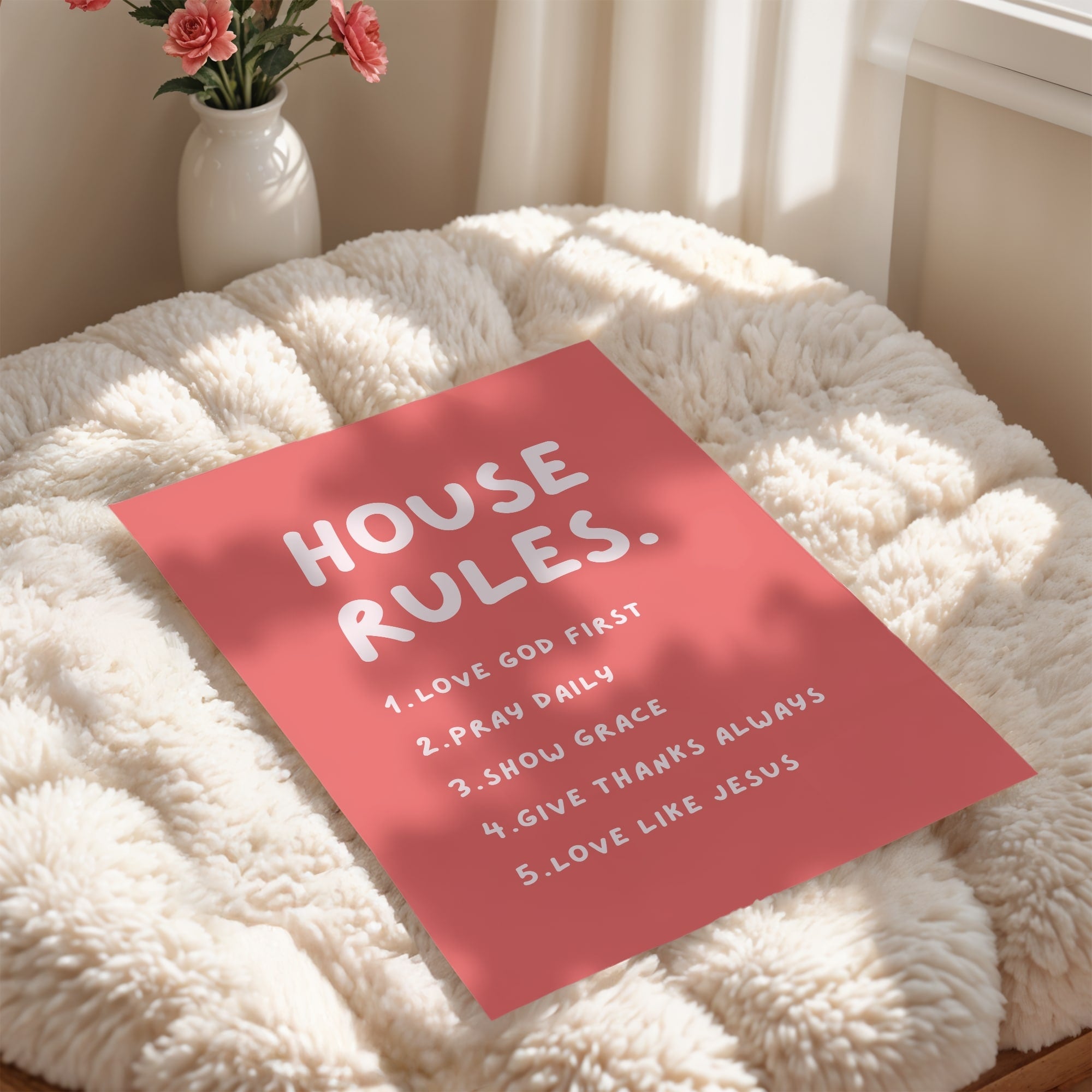 Coral unframed poster with faith filledd 'House Rules' on a fluffy white surface, with a vase of flowers in the background.