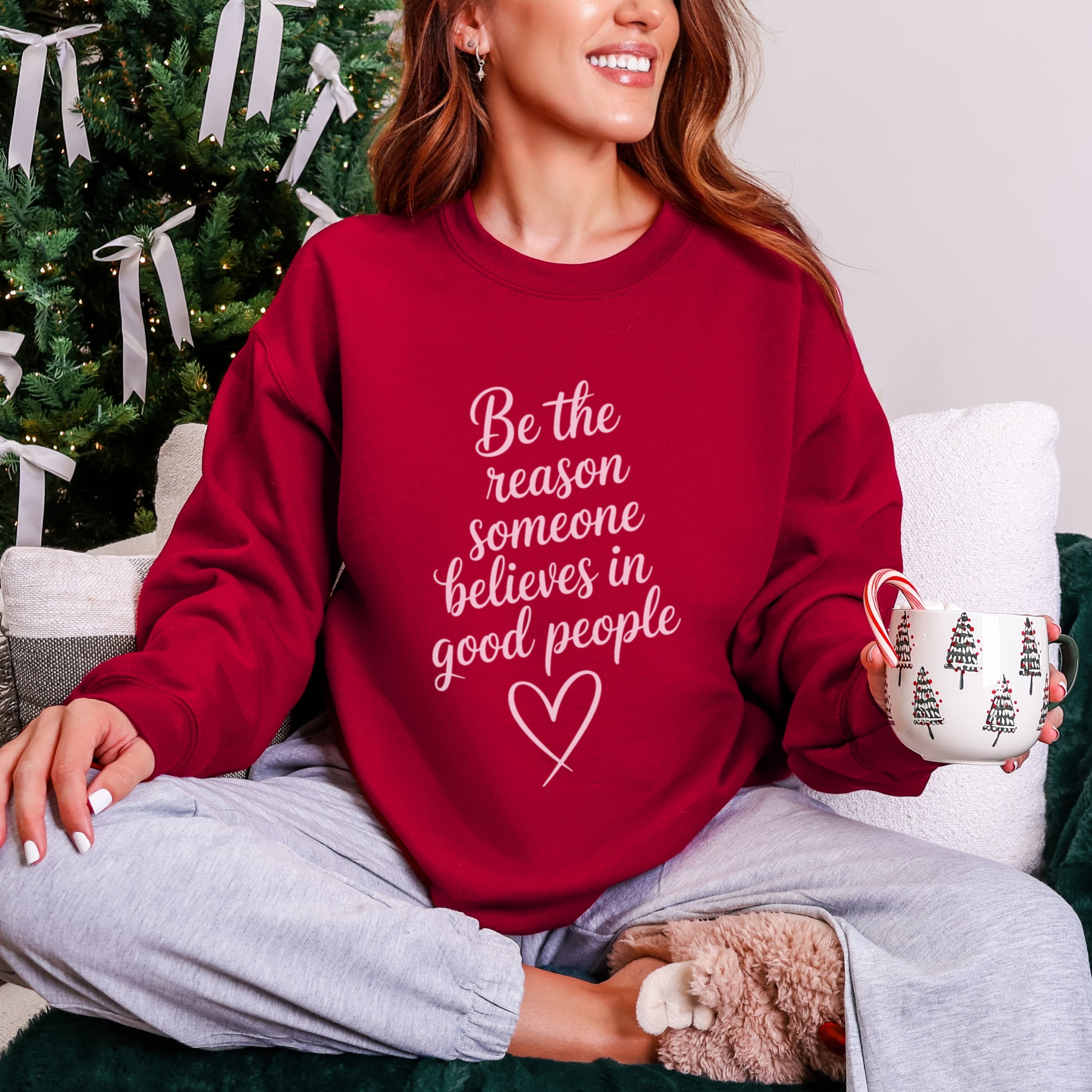 Woman wearing cardinal red Be The Reason Someone Believes In Good People sweatshirt with pale pink text, inspirational kindness crewneck for women in a christmas setting
