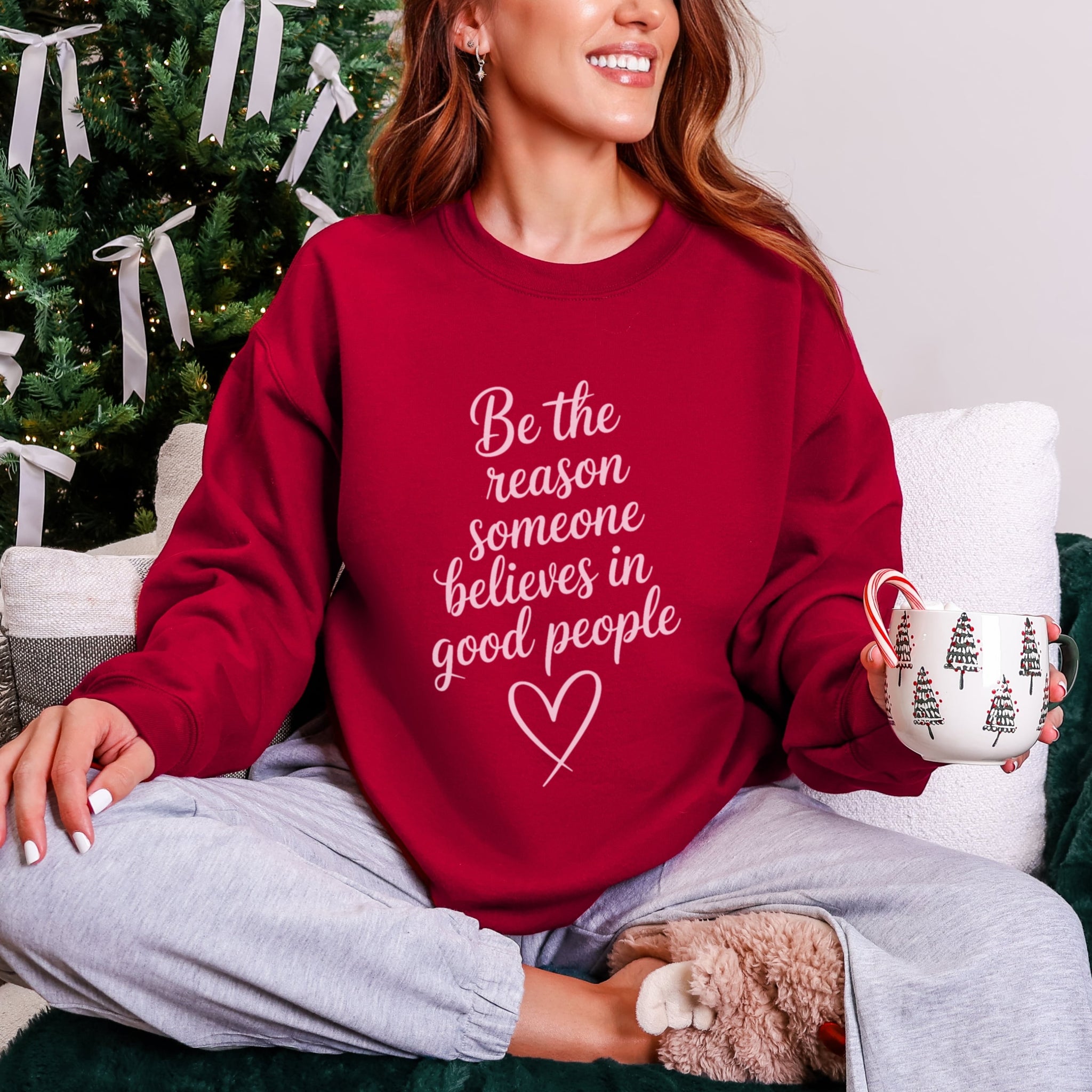 Woman wearing cardinal red Be The Reason Someone Believes In Good People sweatshirt with pale pink text, inspirational kindness crewneck for women in a christmas setting