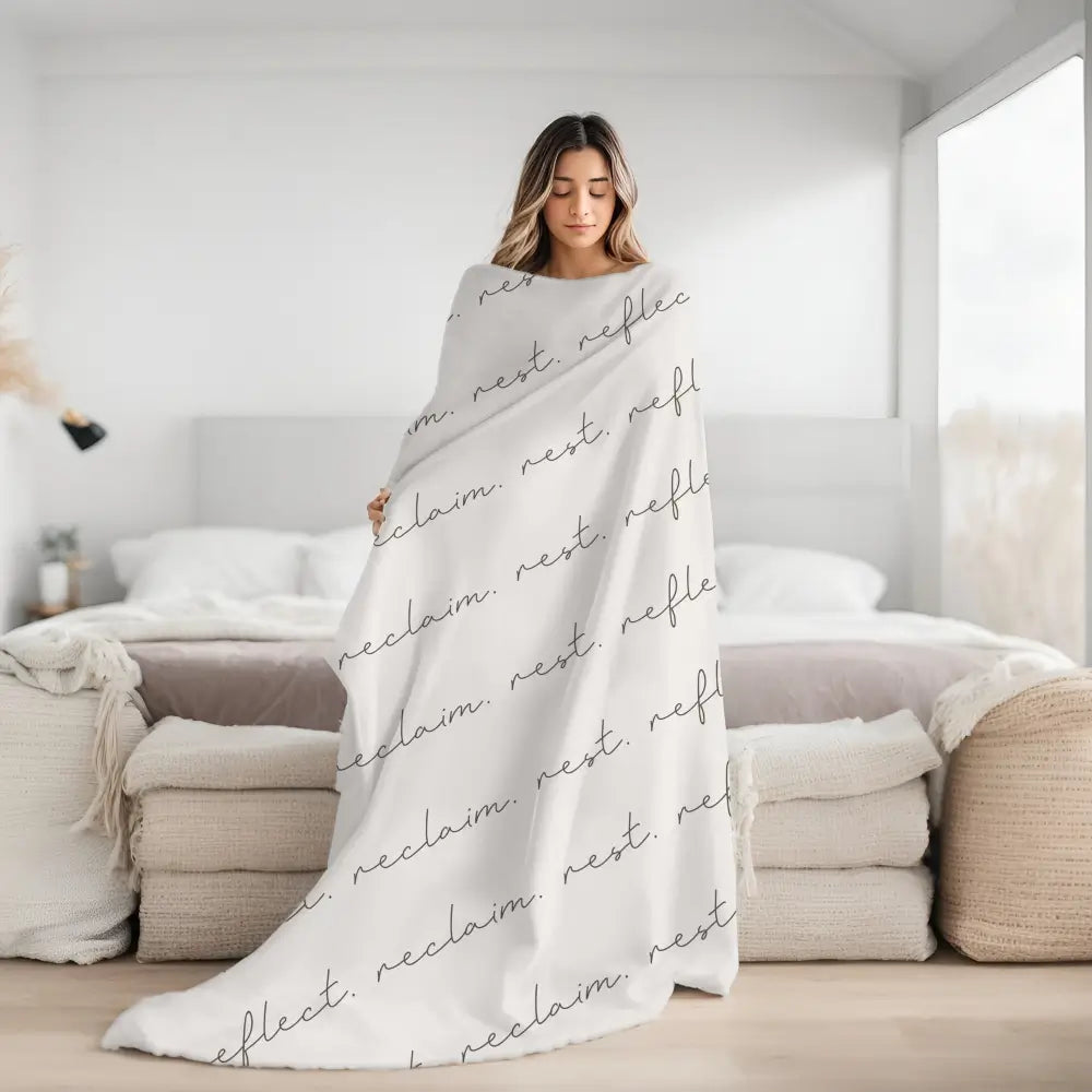 Cozy neutral colored sherpa blanket with repeating reflect. reclaim. rest. in elegant script wrapped around a woman showing its large size