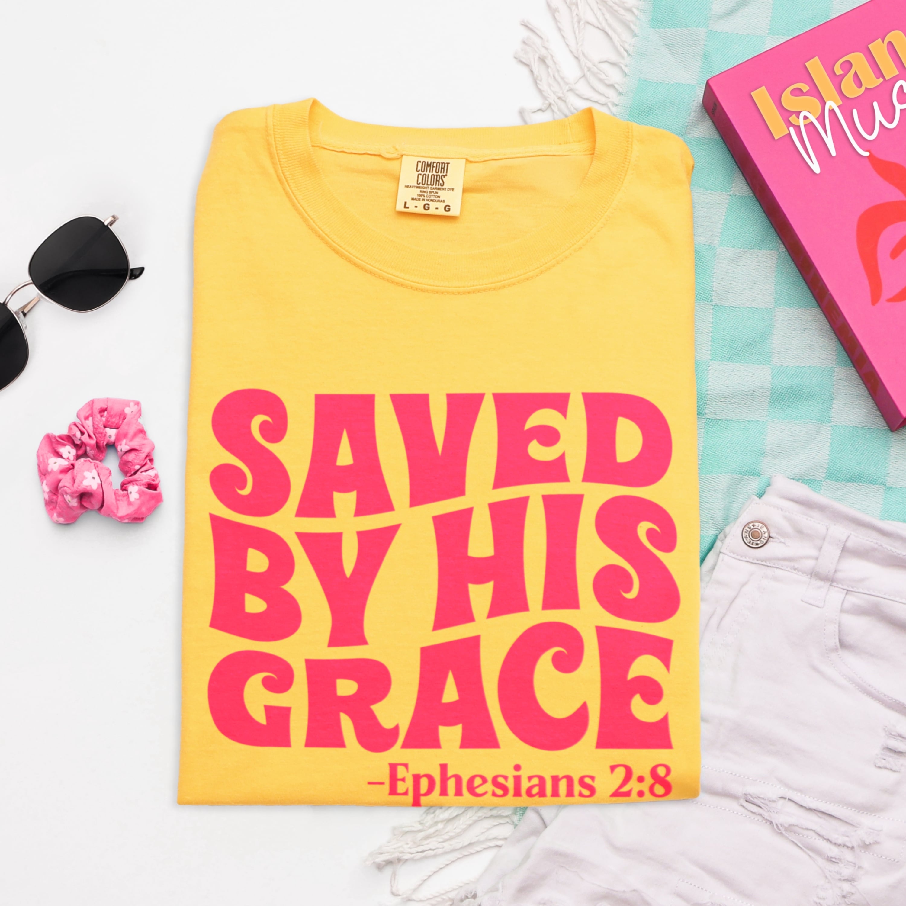 Close up of Saved By His Grace bold design with Ephesians 2:8 scripture in hot pink on yellow cotton tshirt