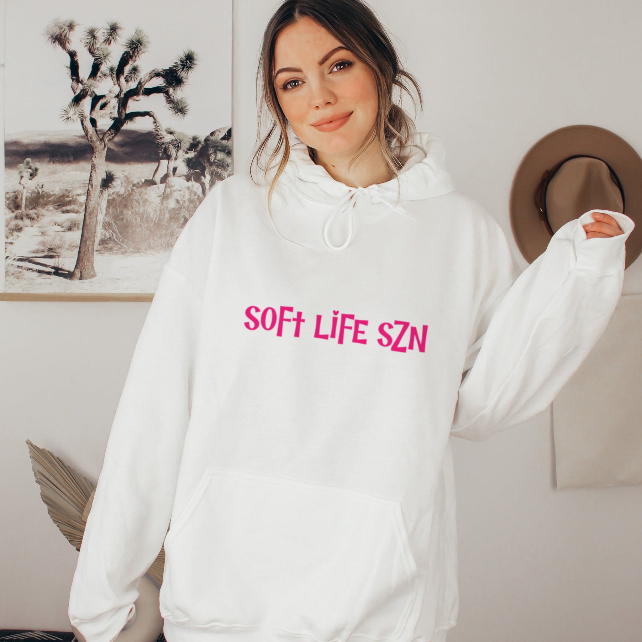 Person wearing a white hoodie with 'SOFT LIFE SZN' text in pink, standing indoors.