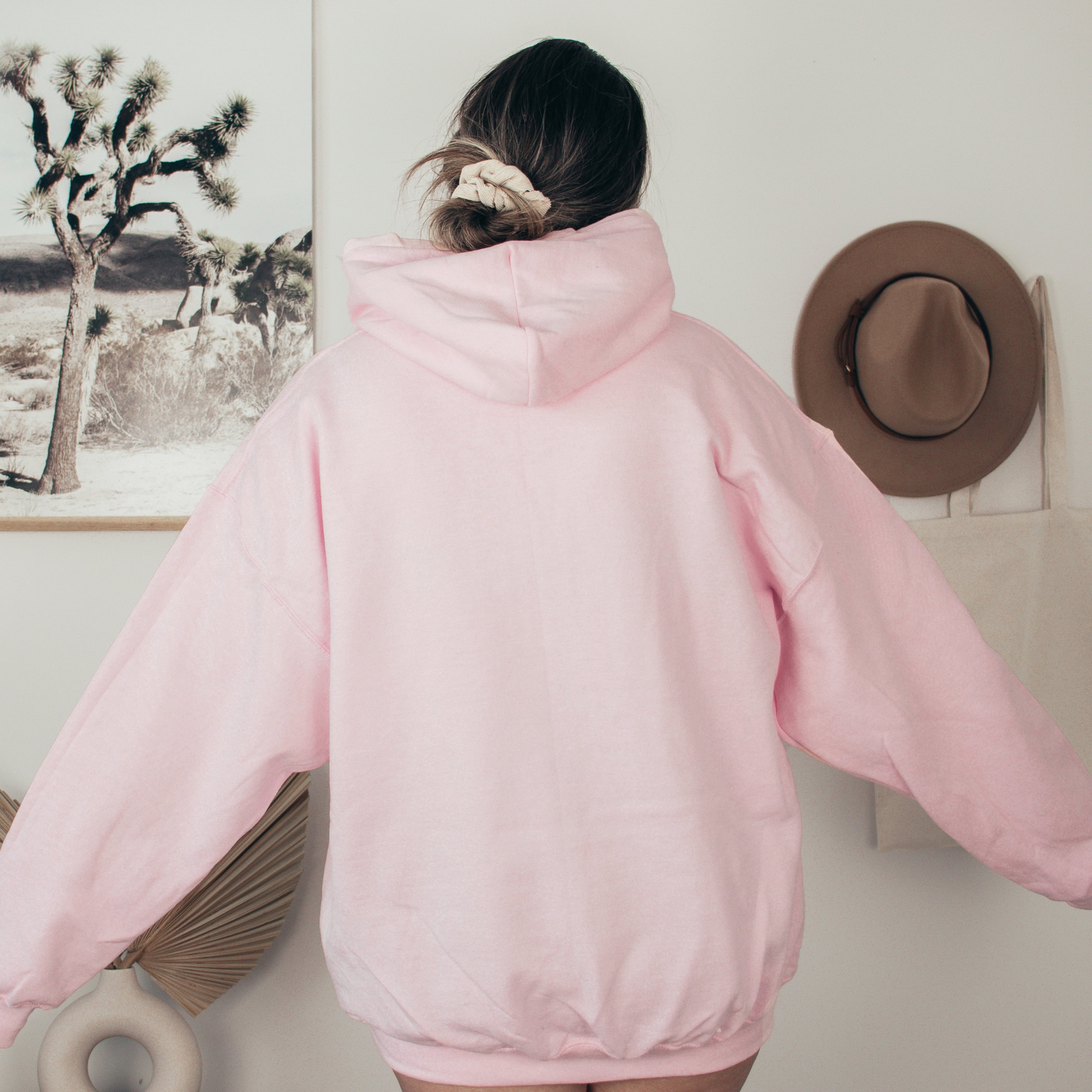 Person wearing a pink hoodie in a room with a hat and artwork on the wall.