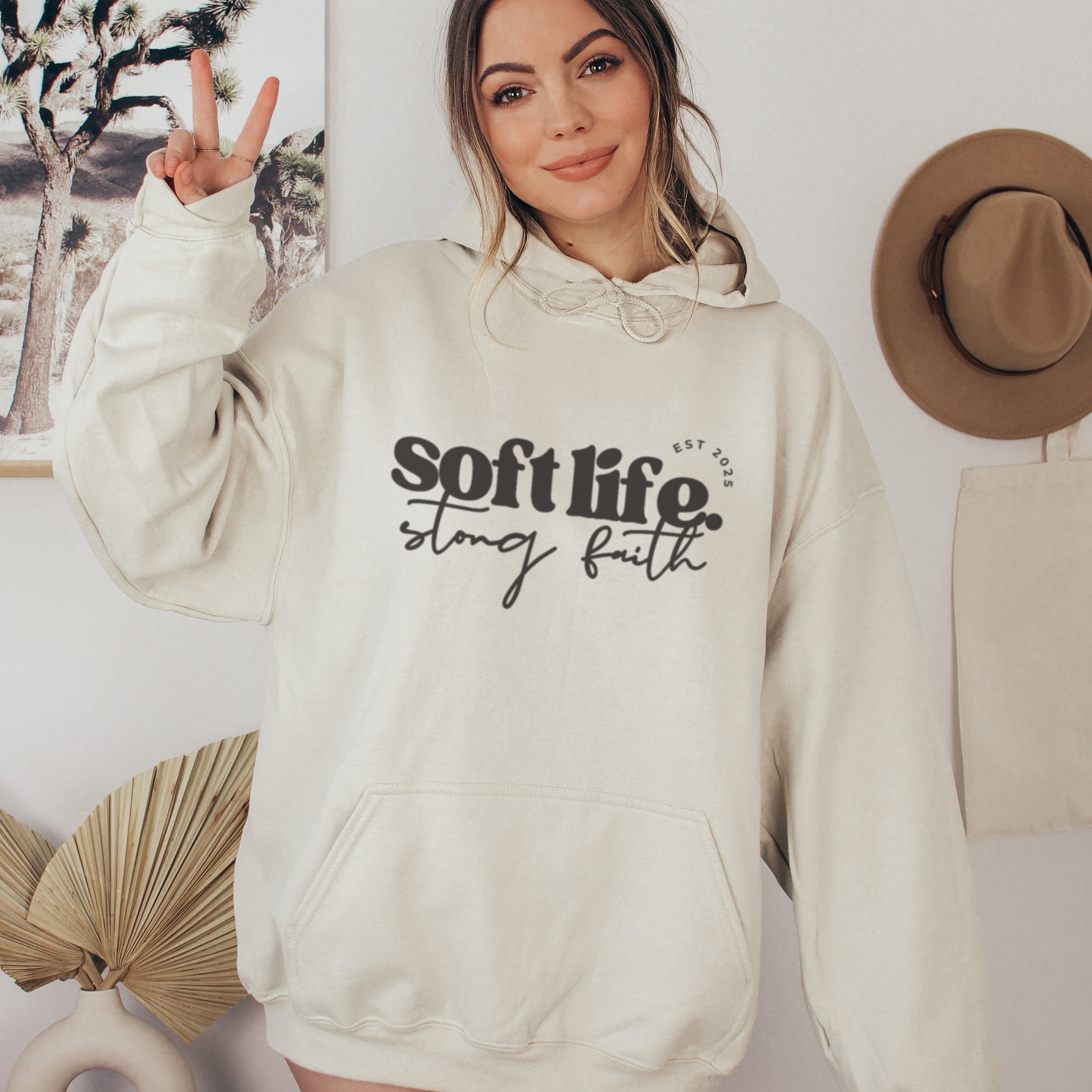 Person wearing a beige hoodie with 'soft life, strong faith' text in a casual setting.