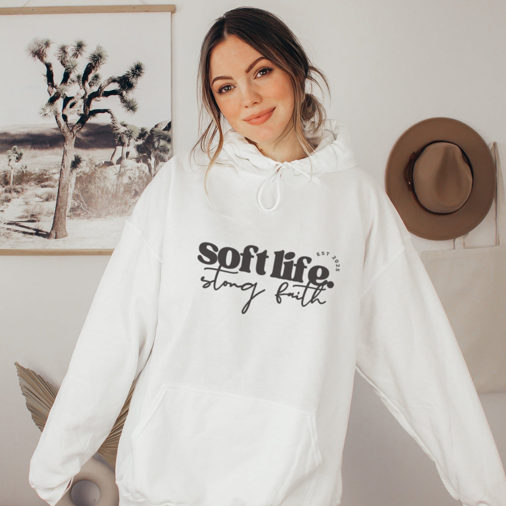 Person wearing a white hoodie with 'soft life, strong faith' text in a room with a hat and artwork on the wall.
