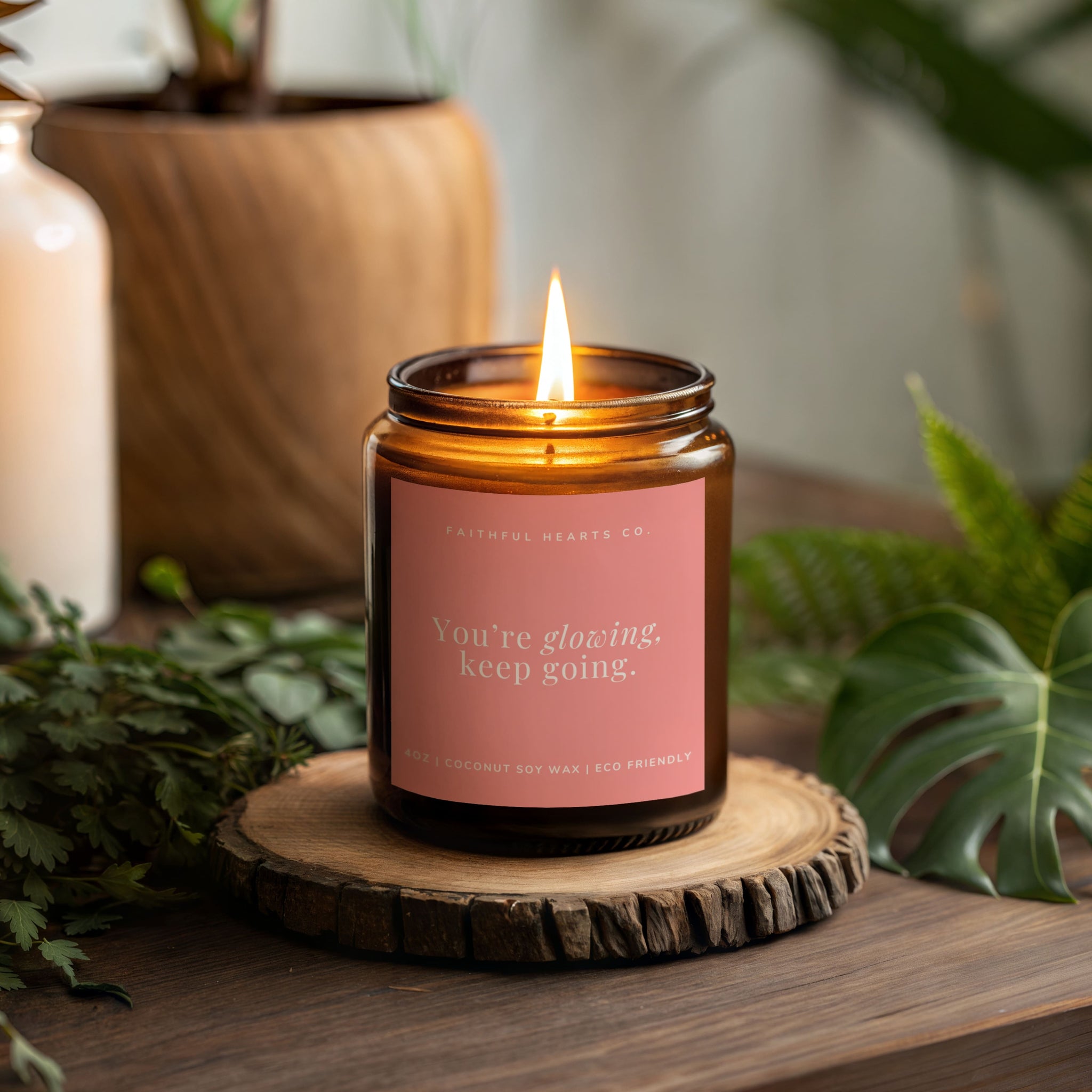 Candle in an amber jar with a pink label on a wooden coaster, surrounded by greenery.