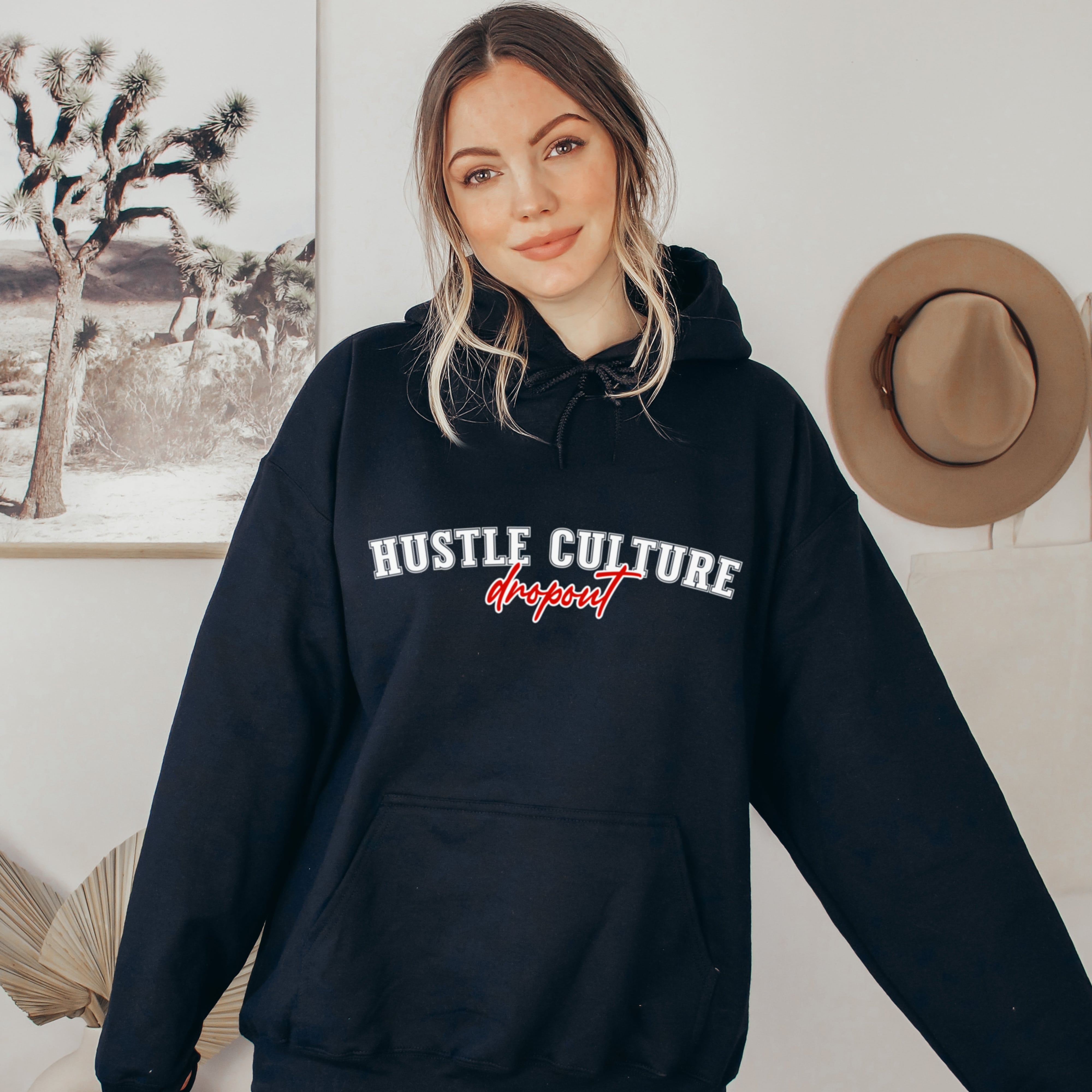 Woman wearing black hustle culture dropout hoodie with white text and red script, soft life anti-hustle clothing